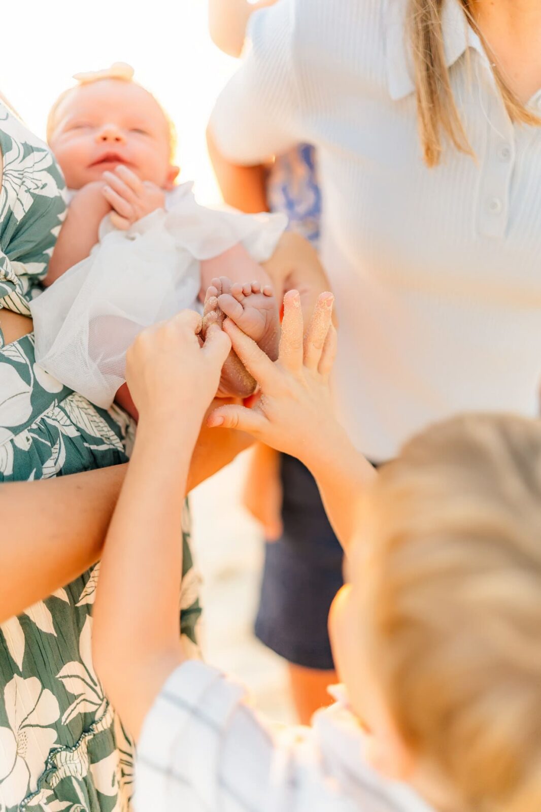 A baby in a white outfit is held by an adult while a child gently touches the baby’s feet. Other adults and children are visible in the sunny, blurred background.