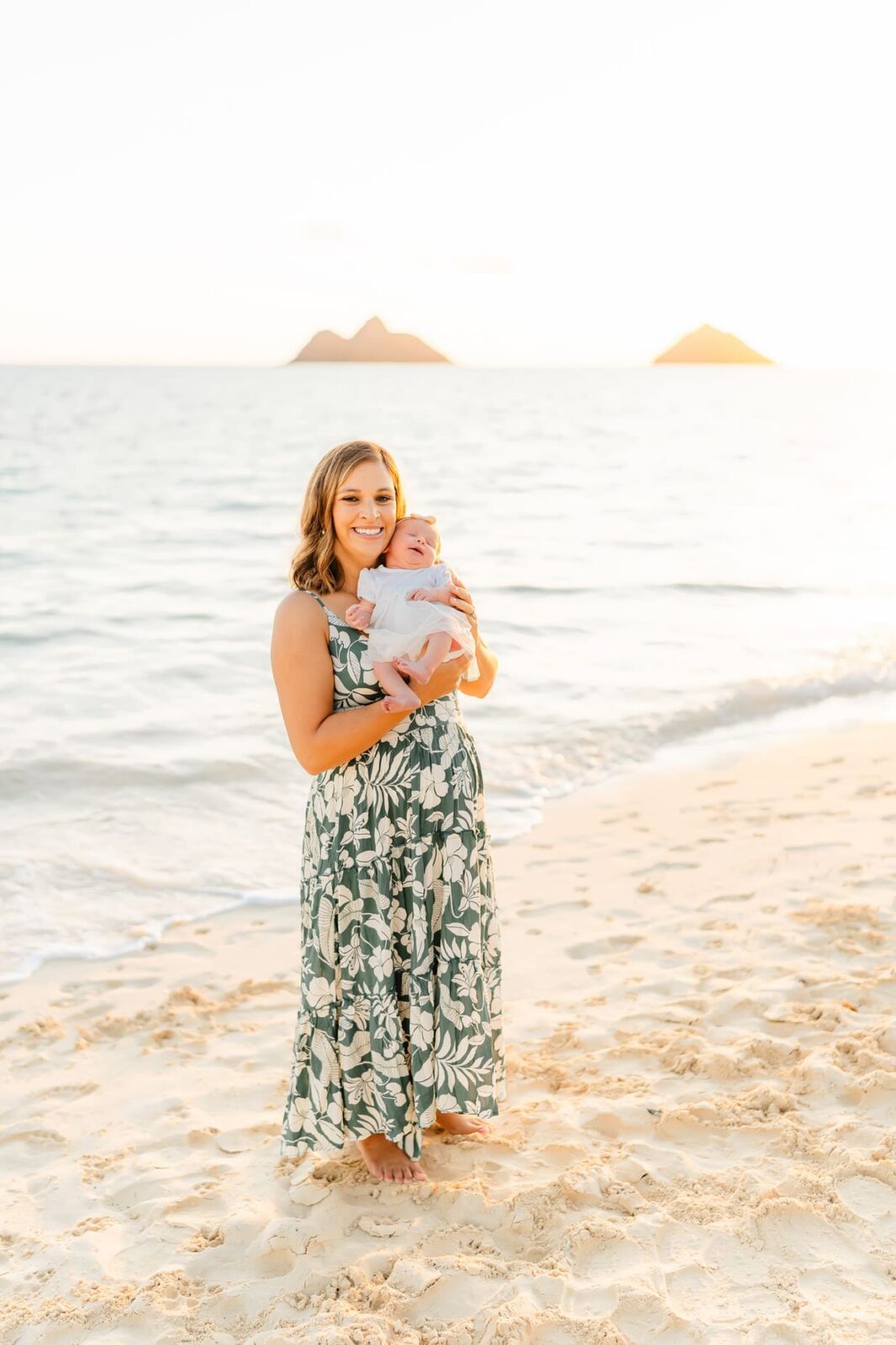 A woman in a long, floral dress stands barefoot on a sandy beach, smiling while holding a baby. The sun is setting over the ocean, with two small islands visible in the background.