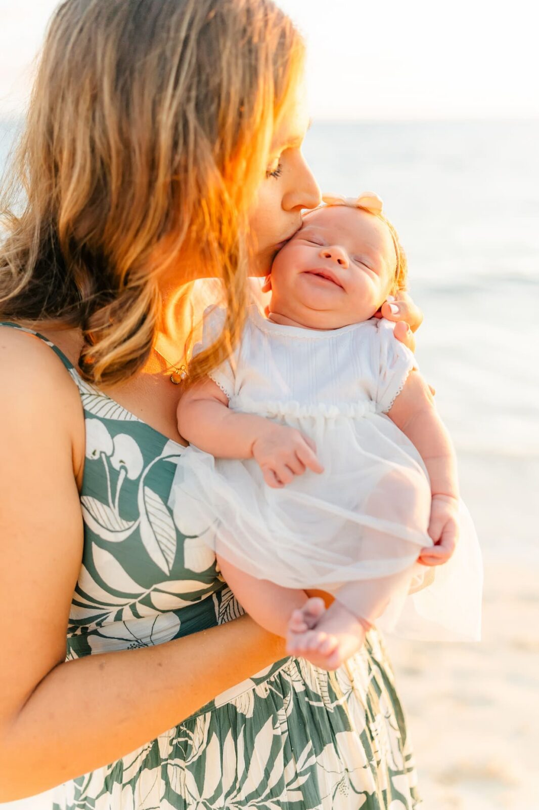 A woman in a floral dress gently kisses a smiling baby’s forehead while holding her at the beach, with soft sunlight illuminating them and the ocean in the background.