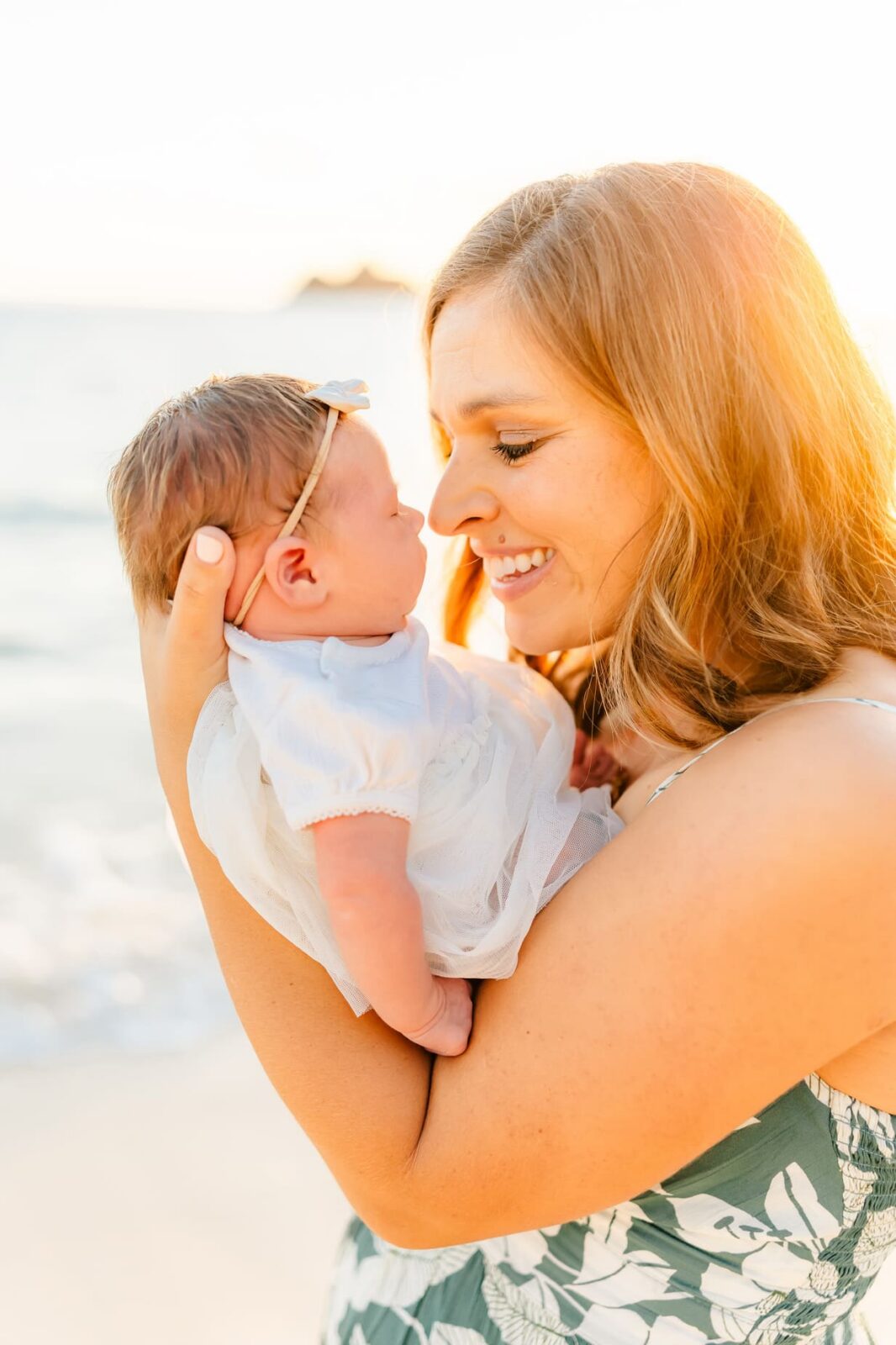 A smiling woman holds a newborn baby close to her face on a sunny beach. The baby wears a white dress and a headband. The background is bright and softly blurred, suggesting a warm, serene atmosphere.