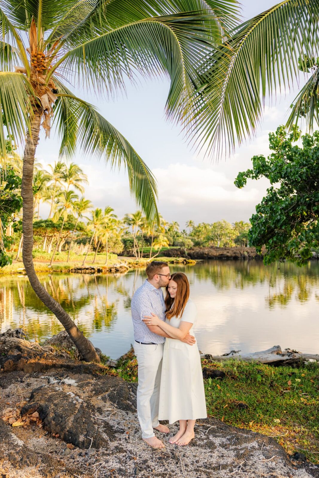 A couple embraces and kisses under palm trees by a calm lake, surrounded by lush greenery and tropical scenery. The sunlight casts a warm glow over the peaceful setting.
