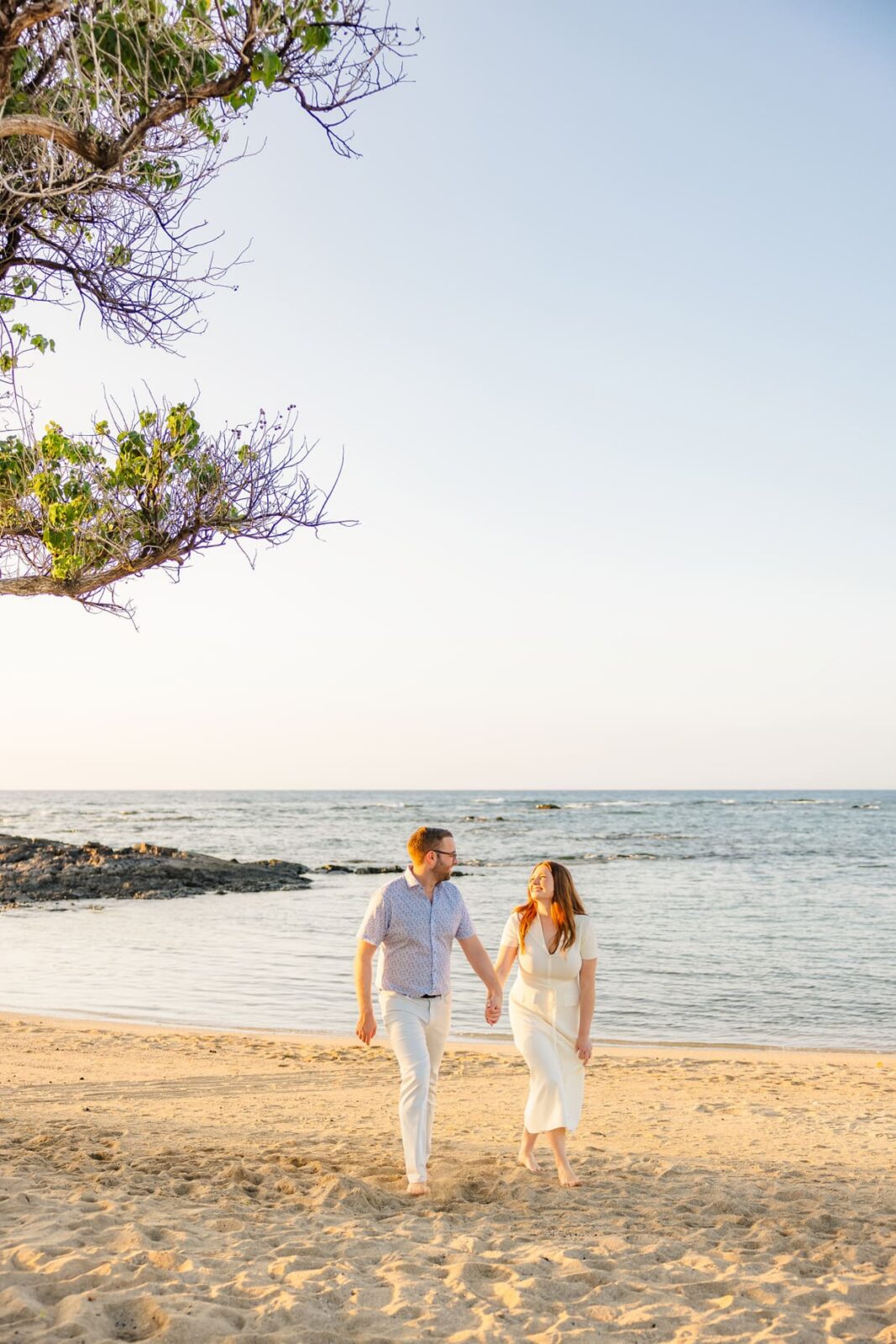 A couple walks barefoot on a sandy beach near the ocean, holding hands and smiling at each other. The sky is clear, and a tree branch extends into the top left corner of the image.