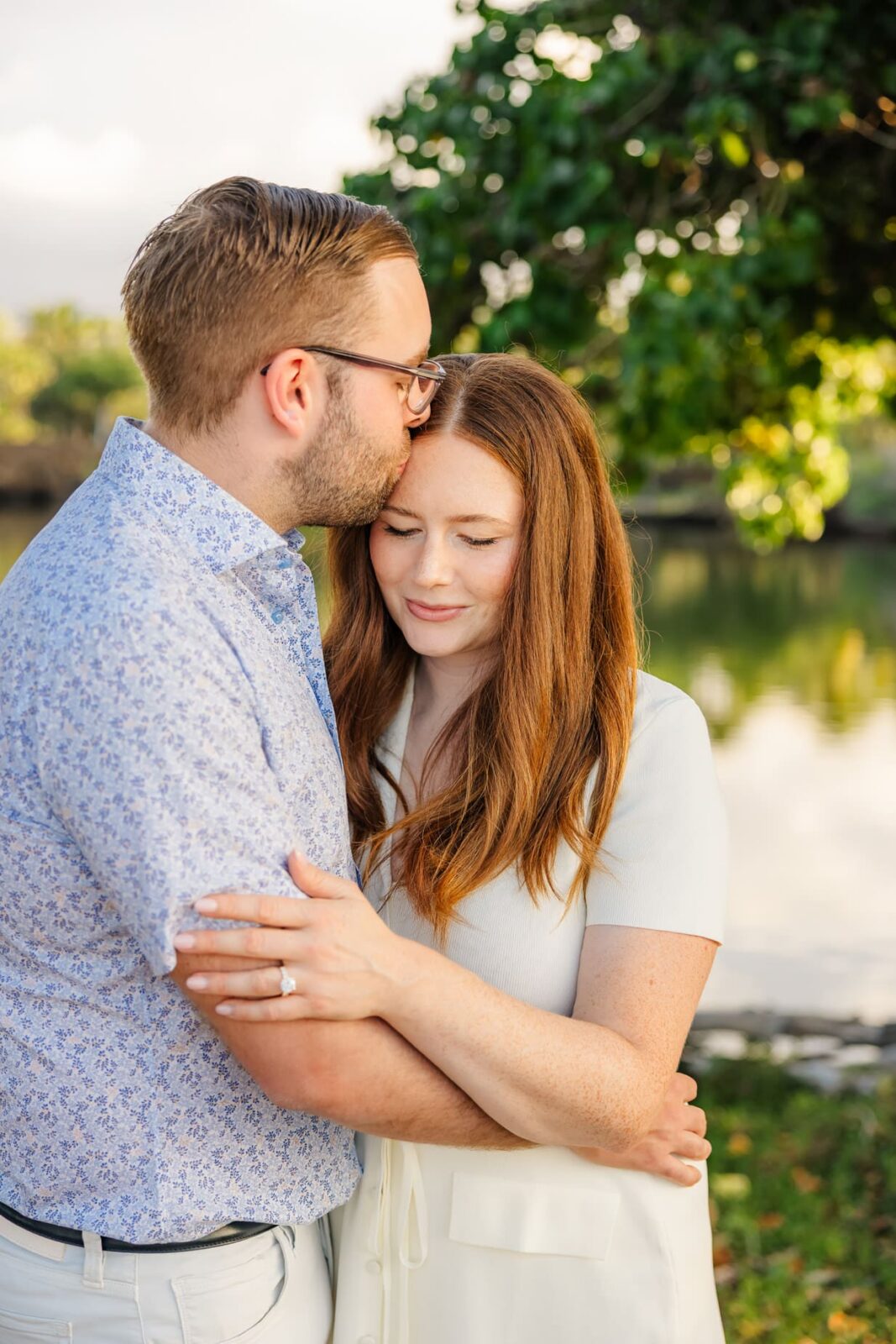 A man kisses a woman on the forehead as they embrace outdoors by a calm lake, both appearing content. The woman gently rests her hand, showing an engagement ring, on the mans arm. Green trees and water are in the background.