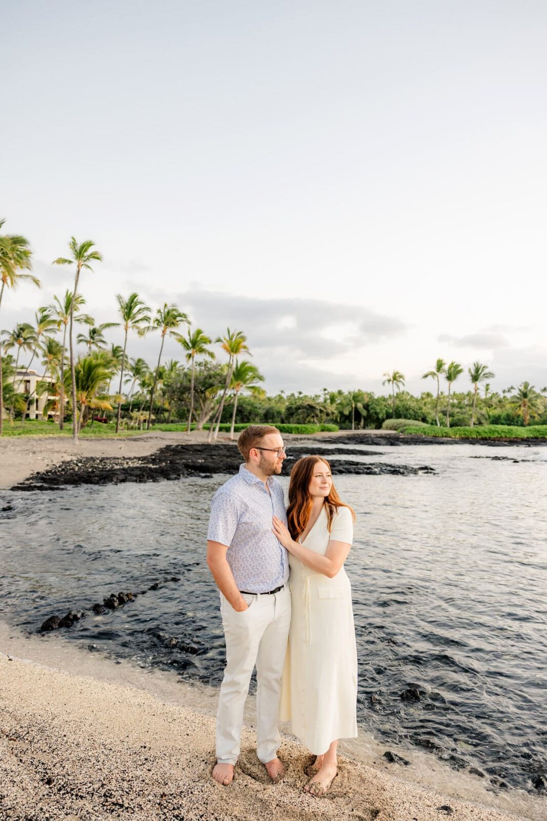 A couple stands barefoot on a sandy beach by the ocean, with palm trees and greenery in the background. The woman rests her hand on the mans chest as they gaze into the distance under a partly cloudy sky.