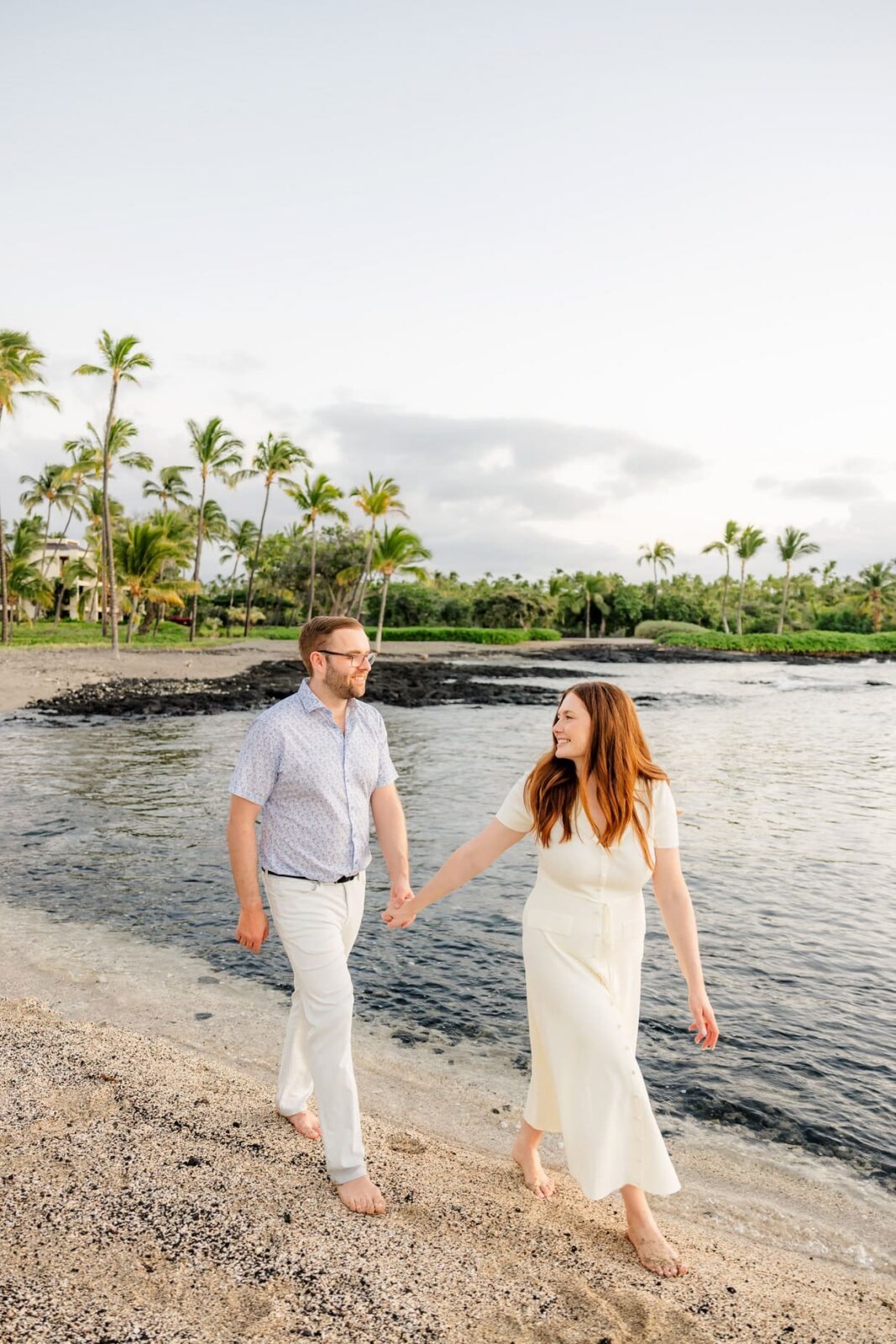 A couple walks barefoot along a sandy beach, holding hands and smiling at each other, with palm trees and calm water in the background under a partly cloudy sky.
