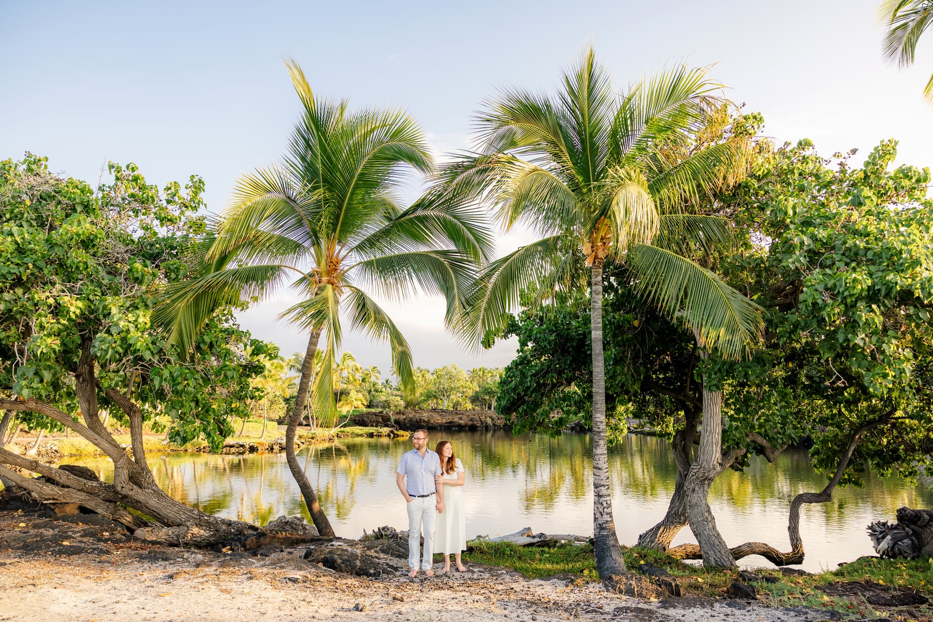A couple in white clothing stands together on a sandy beach, surrounded by palm trees and lush greenery, with a calm body of water and blue sky in the background.