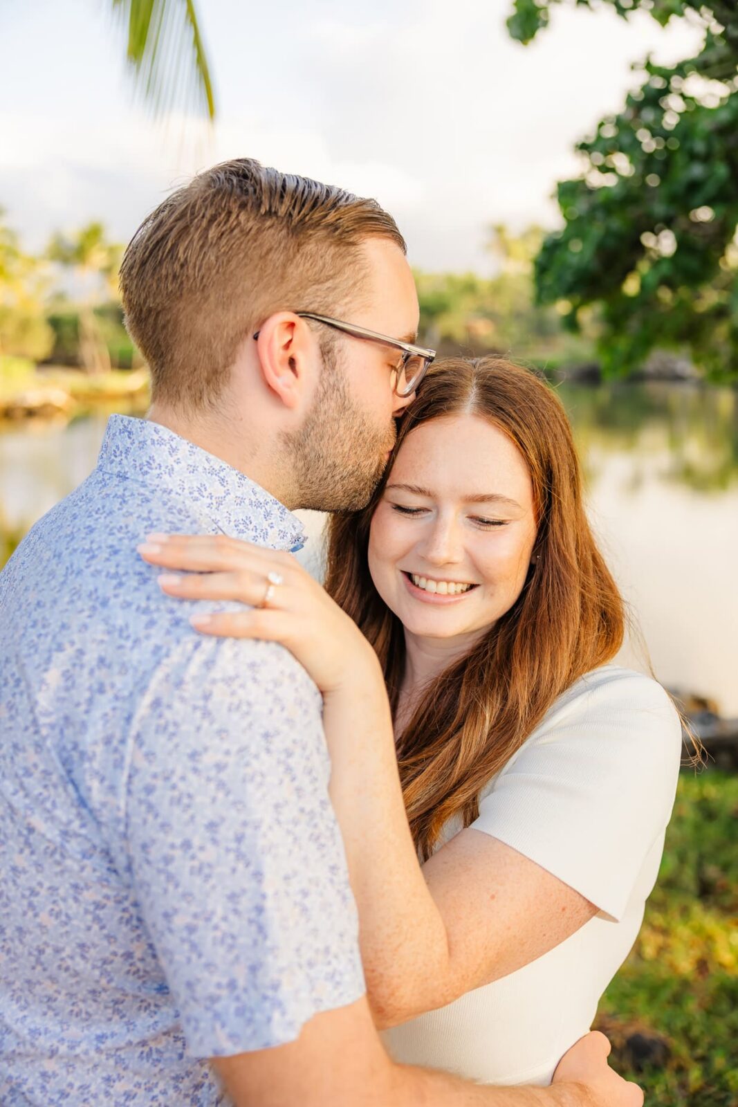 A man kisses a smiling woman on the forehead as they embrace outdoors near a calm body of water, with greenery and palm trees in the background. The woman’s engagement ring is visible on her hand.