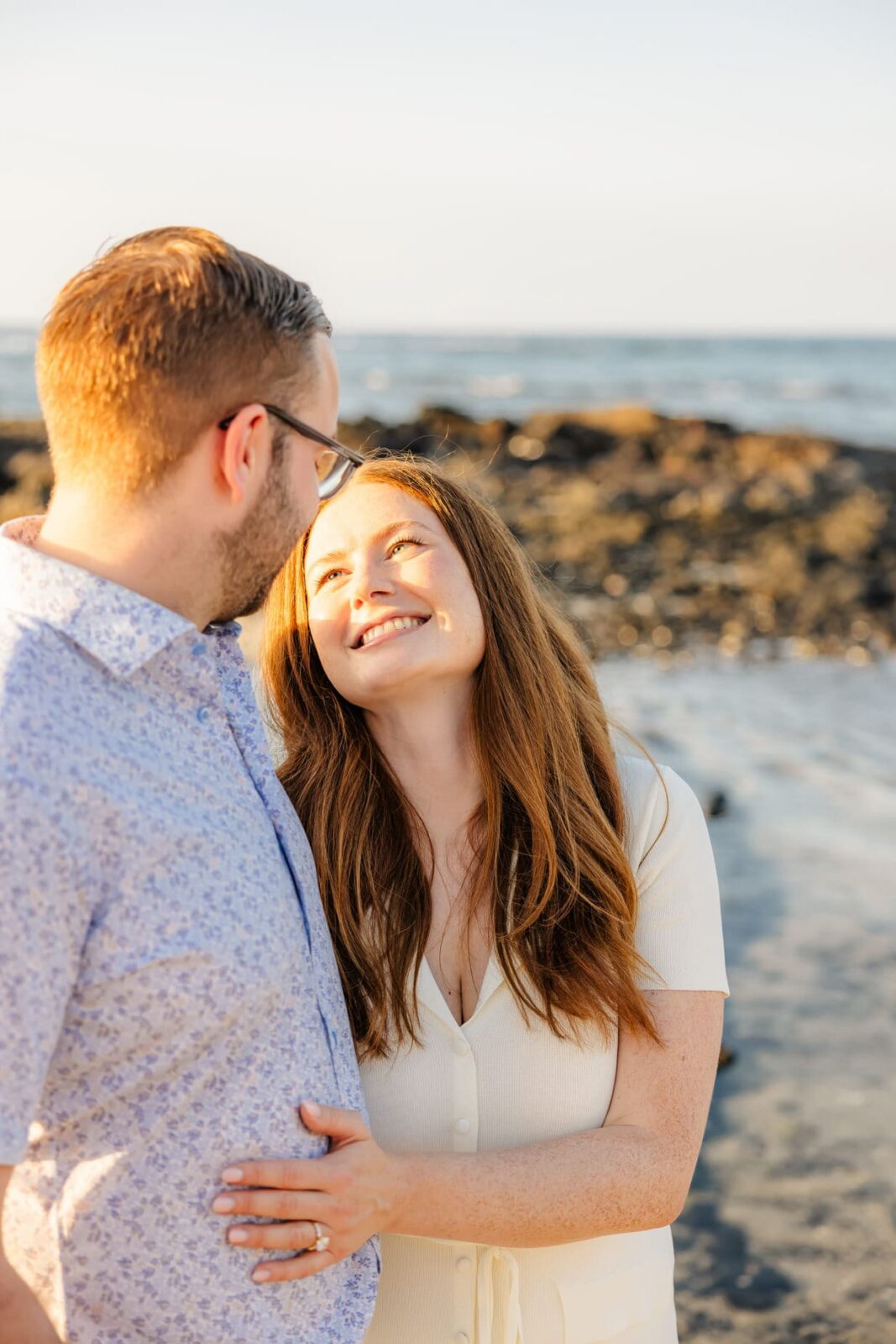 A woman with long red hair smiles up at a man with glasses as they stand close together on a beach, with rocks and the ocean in the background. The sunlight creates a warm, glowing atmosphere.