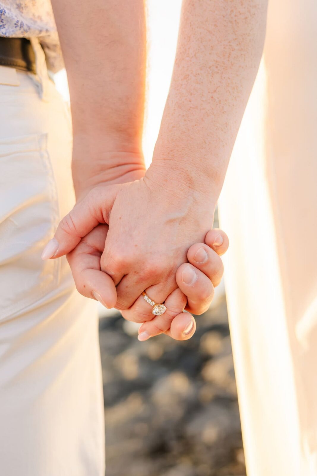 Close-up of two people holding hands, one wearing a diamond engagement ring. They are standing in sunlight, dressed in light-colored clothing, with a blurred outdoor background.