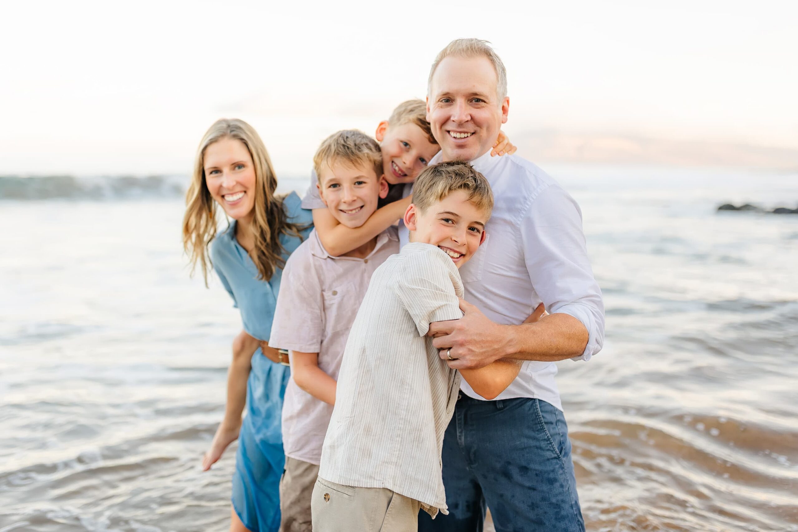 A smiling family of five, with three young boys, stands close together on a beach with gentle waves and a bright sky in the background. The family looks happy and relaxed, wearing light, casual clothing.