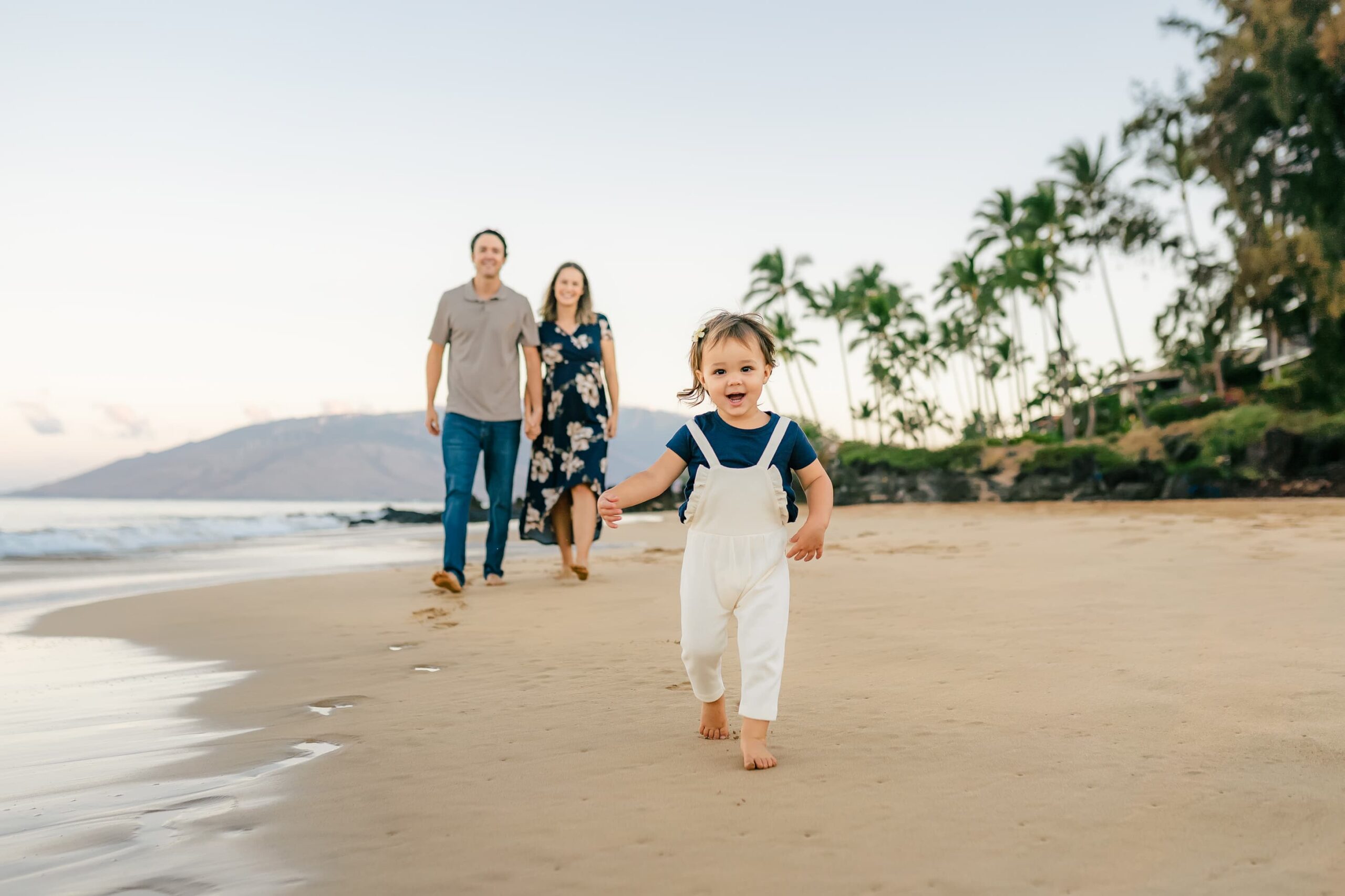 A young child in white overalls runs barefoot on a sandy beach, smiling, with two adults walking behind him. Palm trees and mountains are visible in the background under a clear sky.