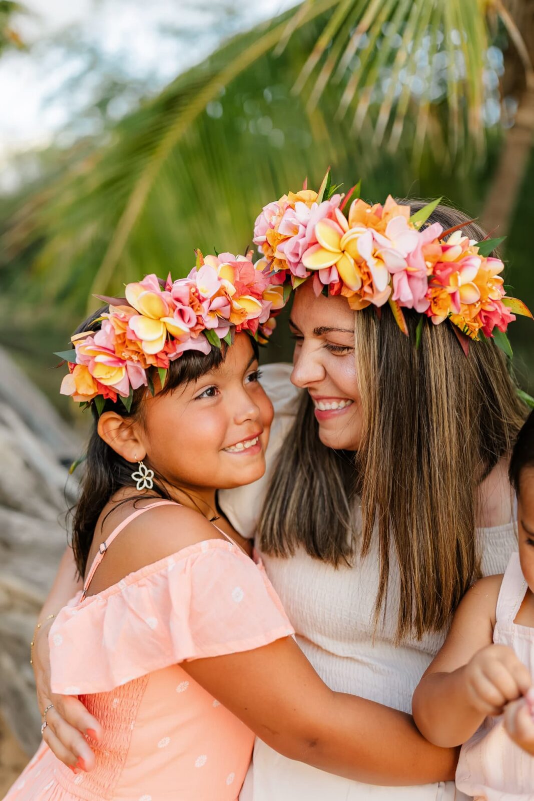 A woman and a young girl wearing colorful flower crowns smile and hug each other outdoors, with palm leaves in the background. Both are dressed in light, summery clothing.