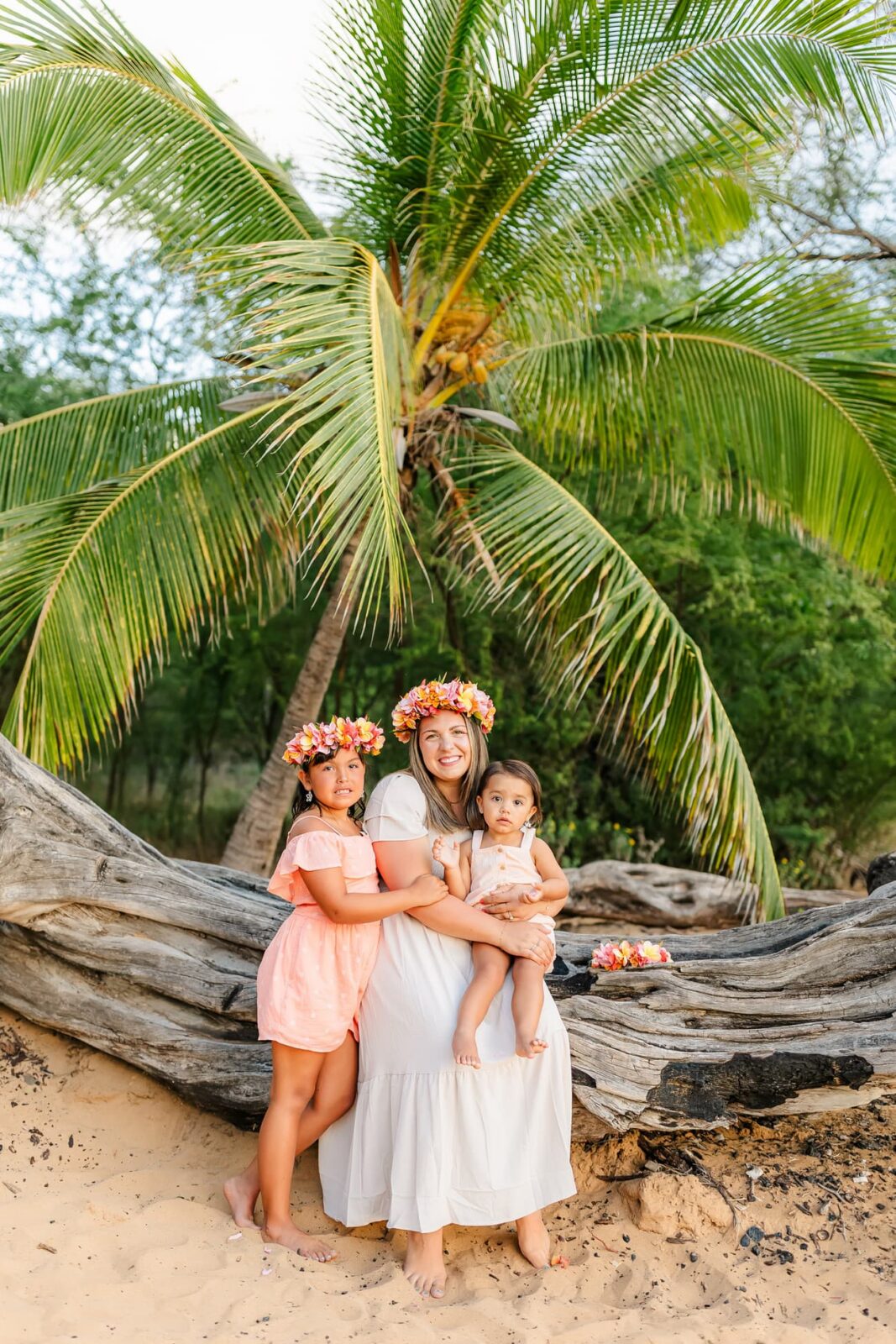 A woman and two young girls, all wearing flower crowns, pose in front of a palm tree and large driftwood on a sandy beach. The woman holds one girl while the other stands beside her in a pink dress.