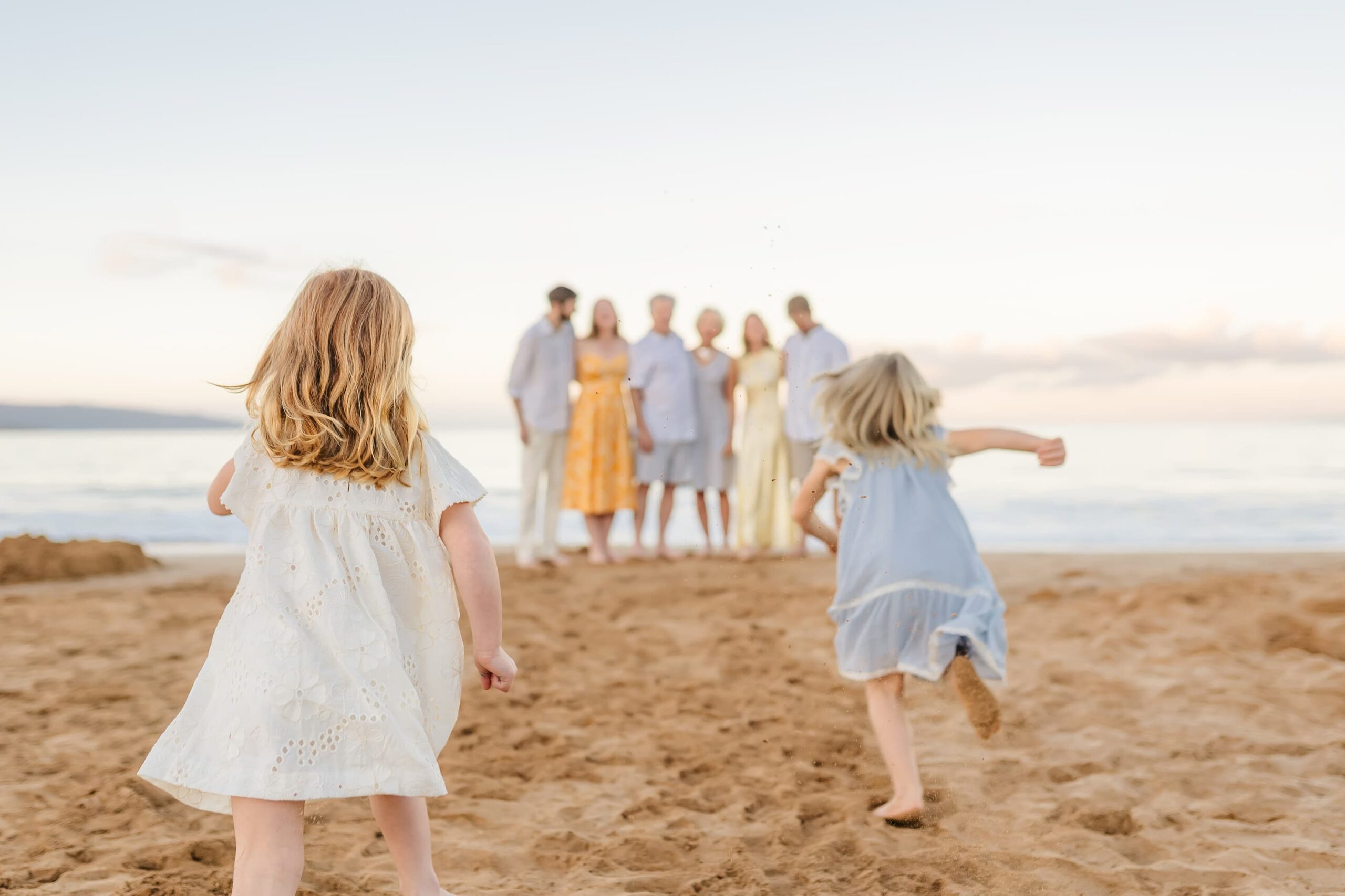 Two young girls in light dresses run on a sandy beach towards a group of adults and children standing together near the shoreline, with the ocean and clear sky in the background.
