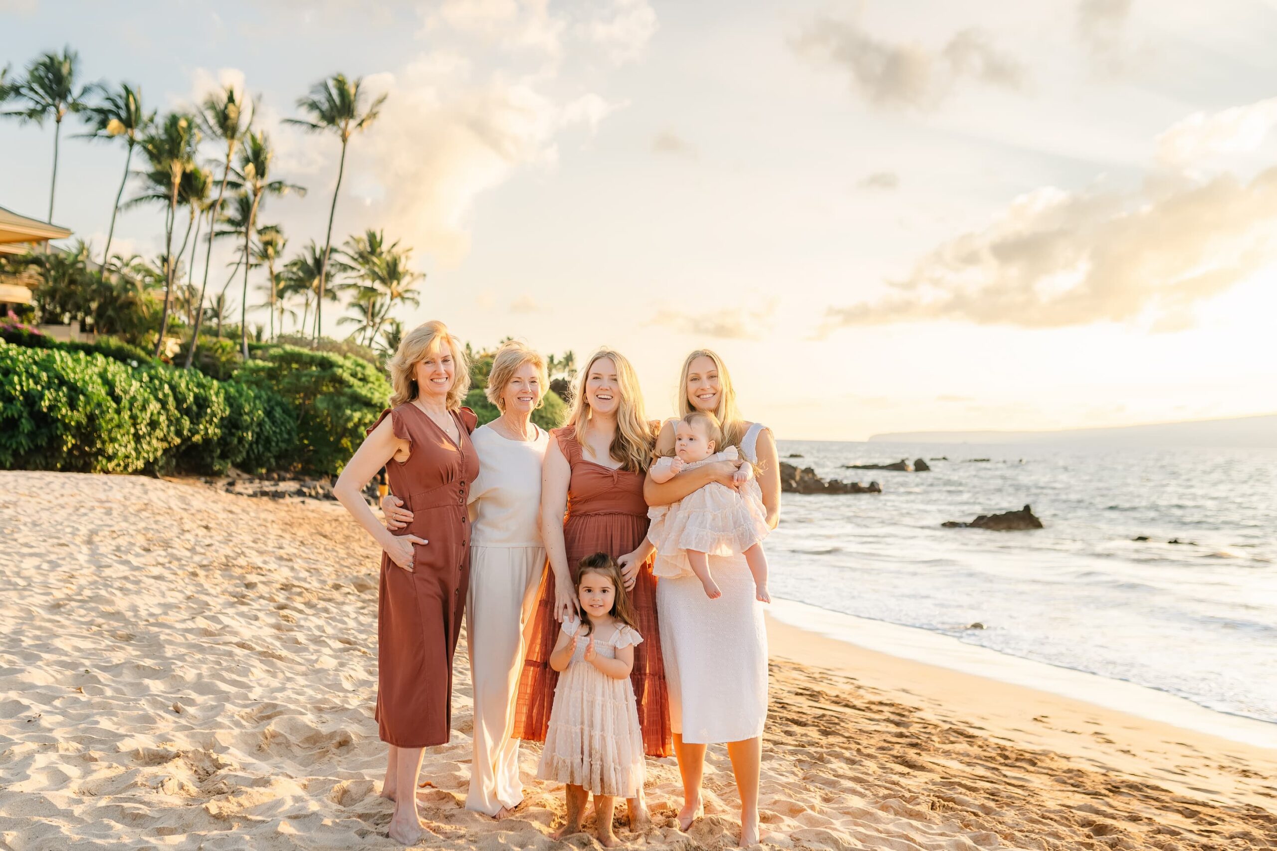 Five women and two young girls stand and smile together on a sandy beach at sunset, with palm trees, greenery, and waves in the background. The group is dressed in light, summery clothing.