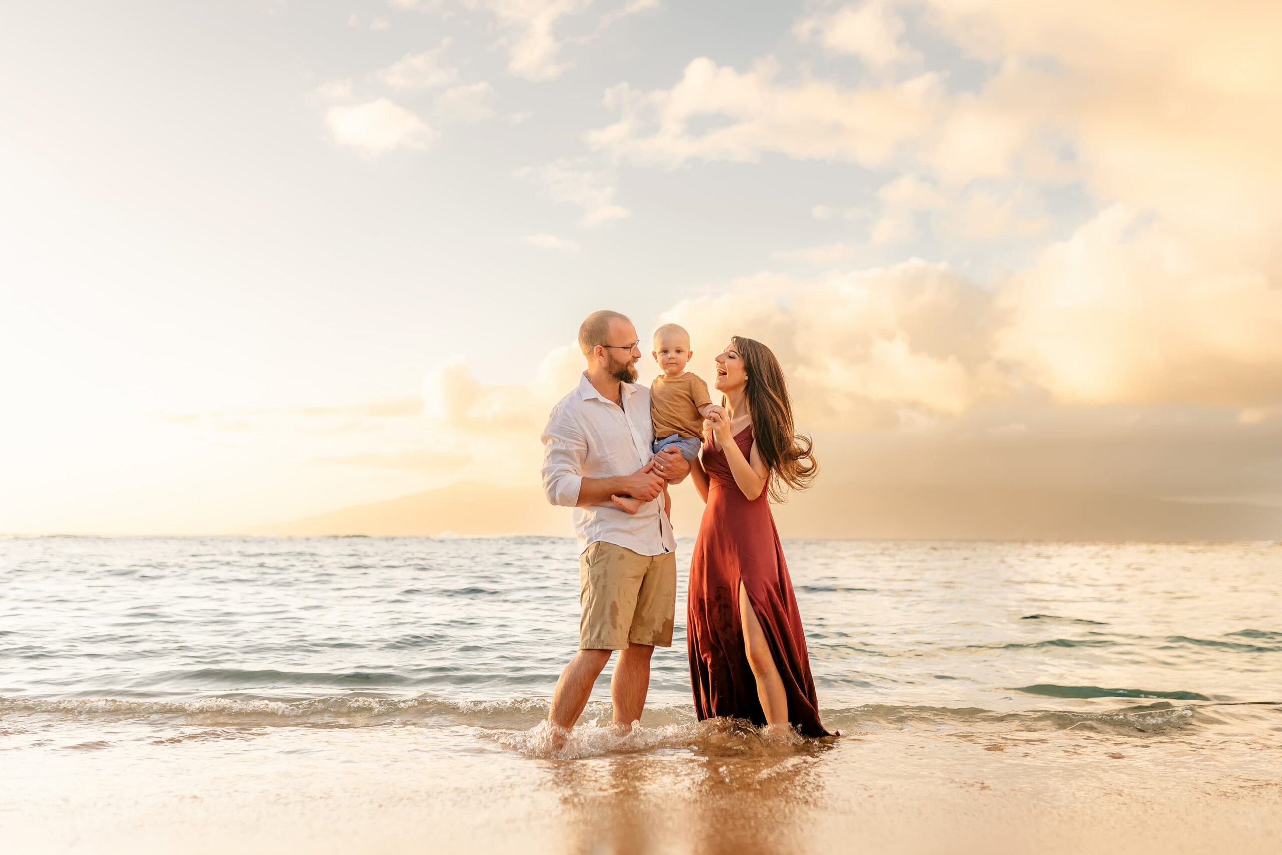 A man and woman stand barefoot in shallow ocean water at sunset, holding a young child between them. The sky is partly cloudy with warm, golden light, and the family appears happy and relaxed.