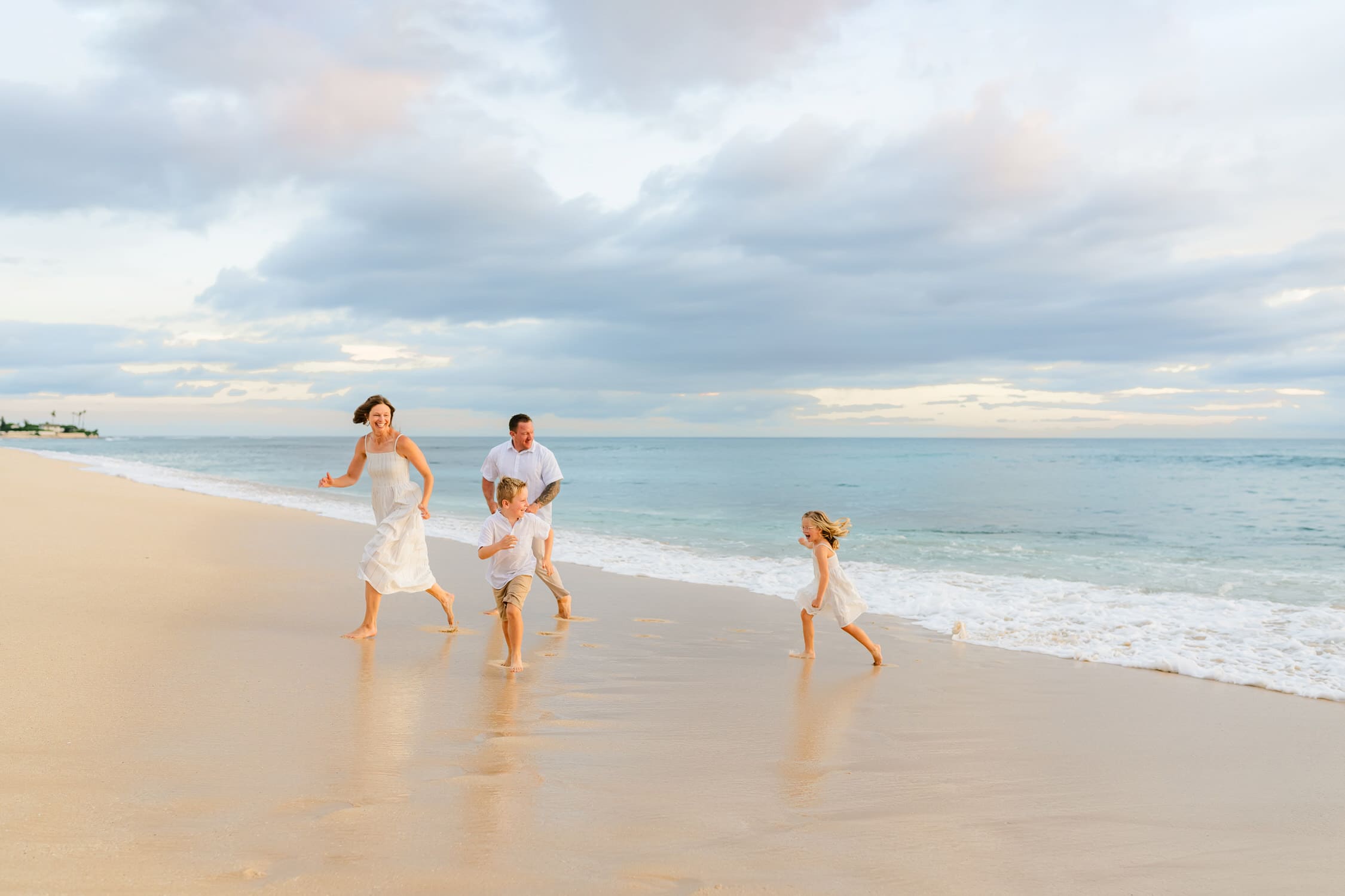 A family of four, dressed in white, runs and plays together on a sandy beach near the ocean under a cloudy sky—capturing unforgettable family photos on your Hawaii vacation.