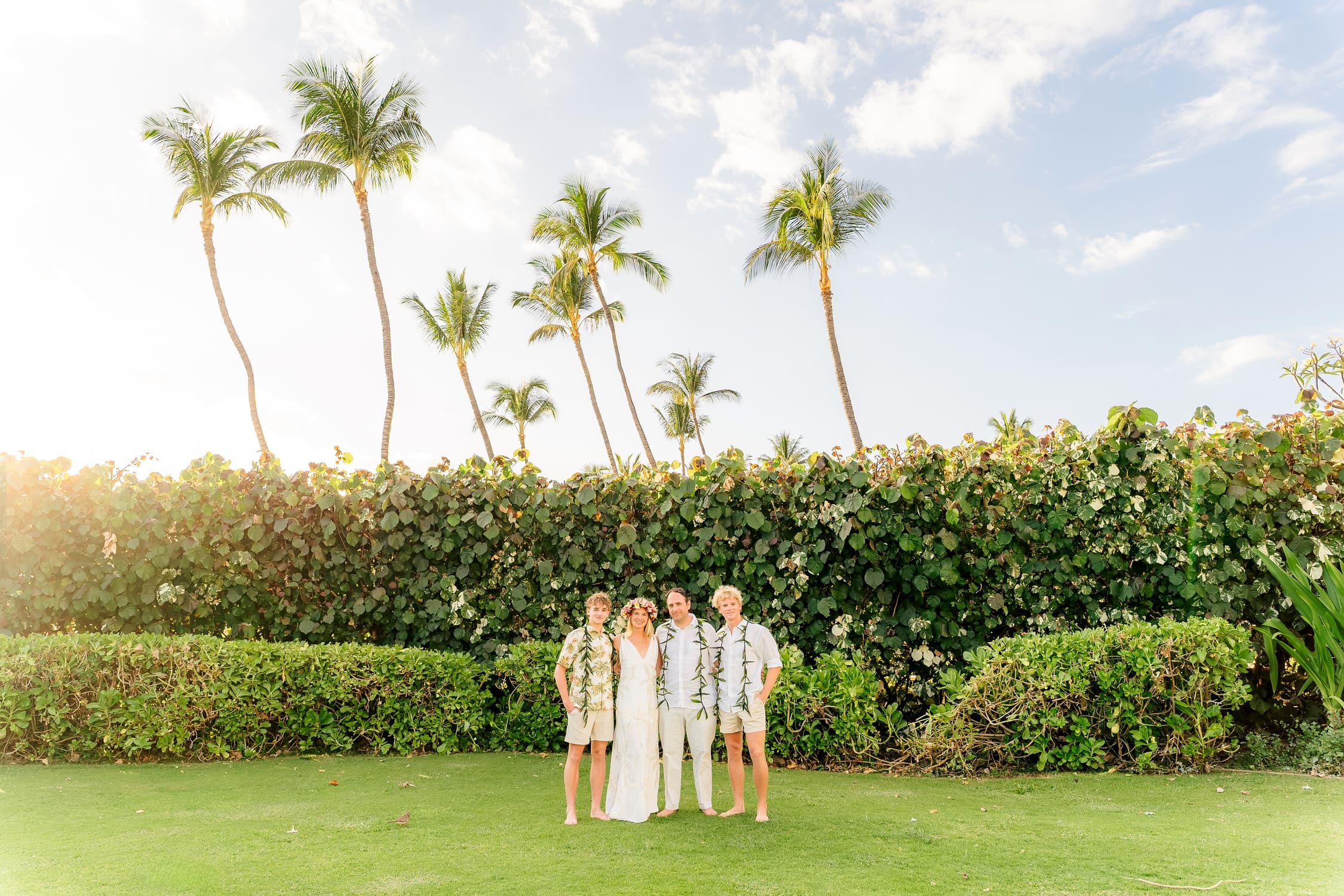 Four people stand together on a green lawn in front of a hedge and tall palm trees under a sunny, blue sky. They are dressed in light, tropical clothing and smiling at the camera.
