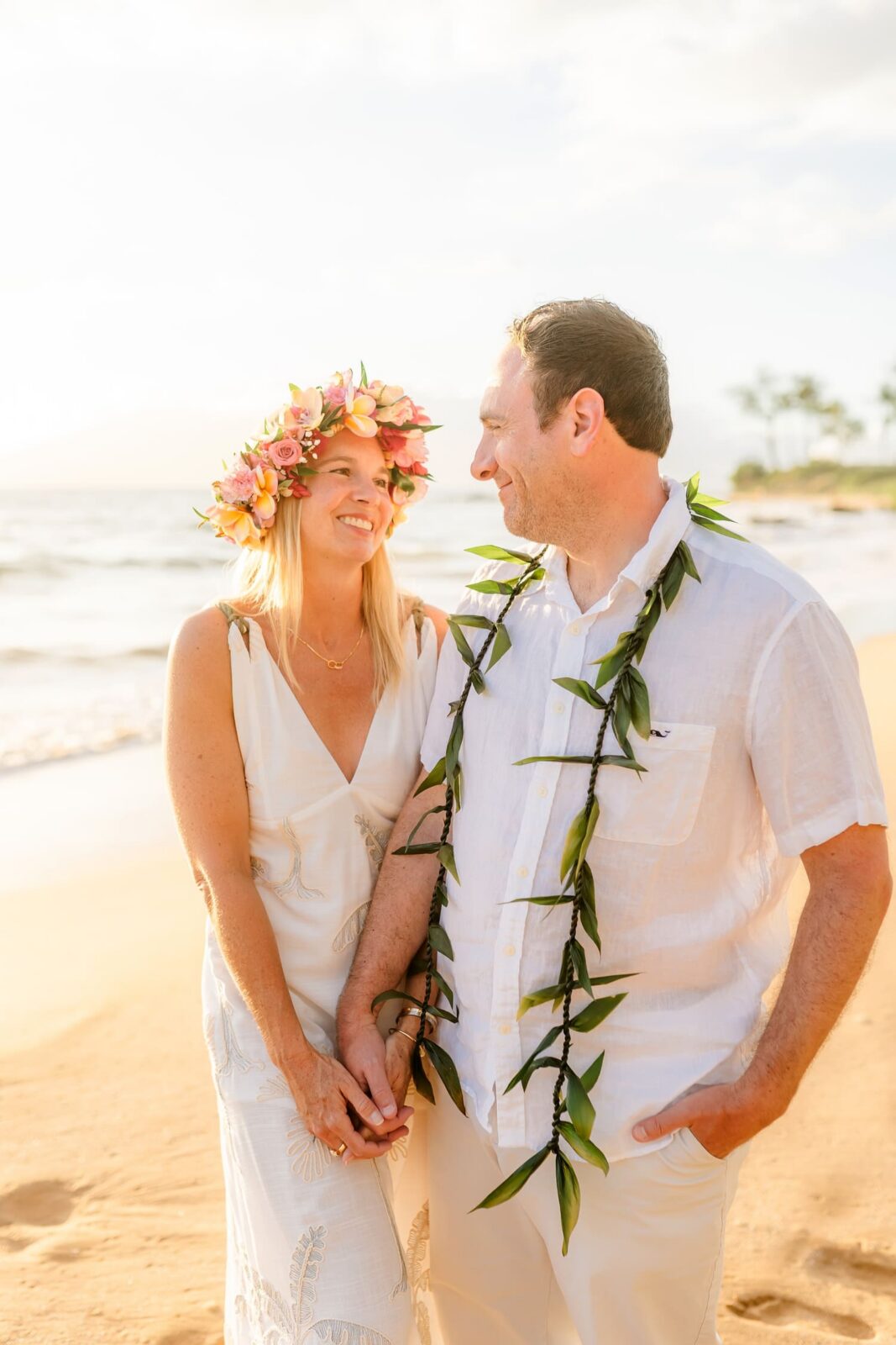 A couple stands on a sunny beach, holding hands and smiling at each other. The woman wears a floral crown and white dress, while the man wears a white shirt with a leafy lei. Waves and palm trees are visible in the background.