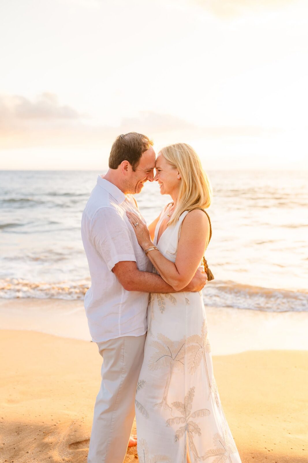 A couple stands on a beach at sunset, embracing and smiling at each other, with the ocean waves and golden sunlight in the background.