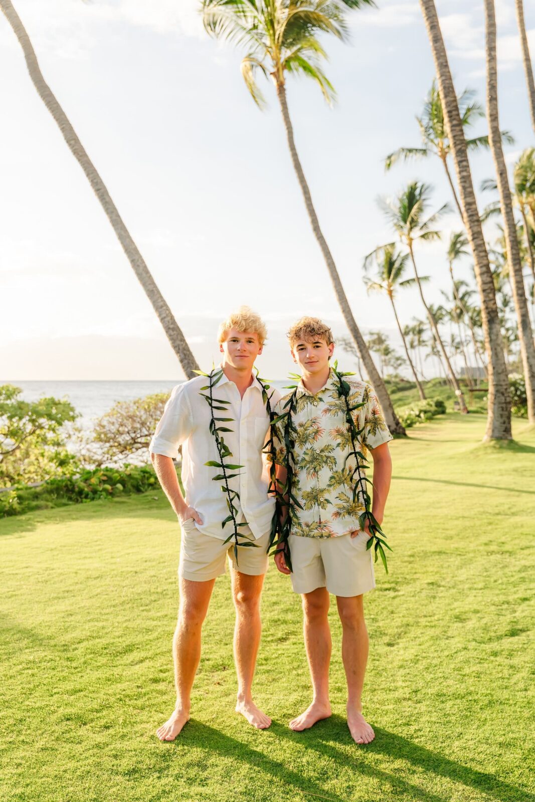 Two young men wearing leis and tropical shirts stand barefoot on green grass with tall palm trees and ocean in the background, under bright sunlight.