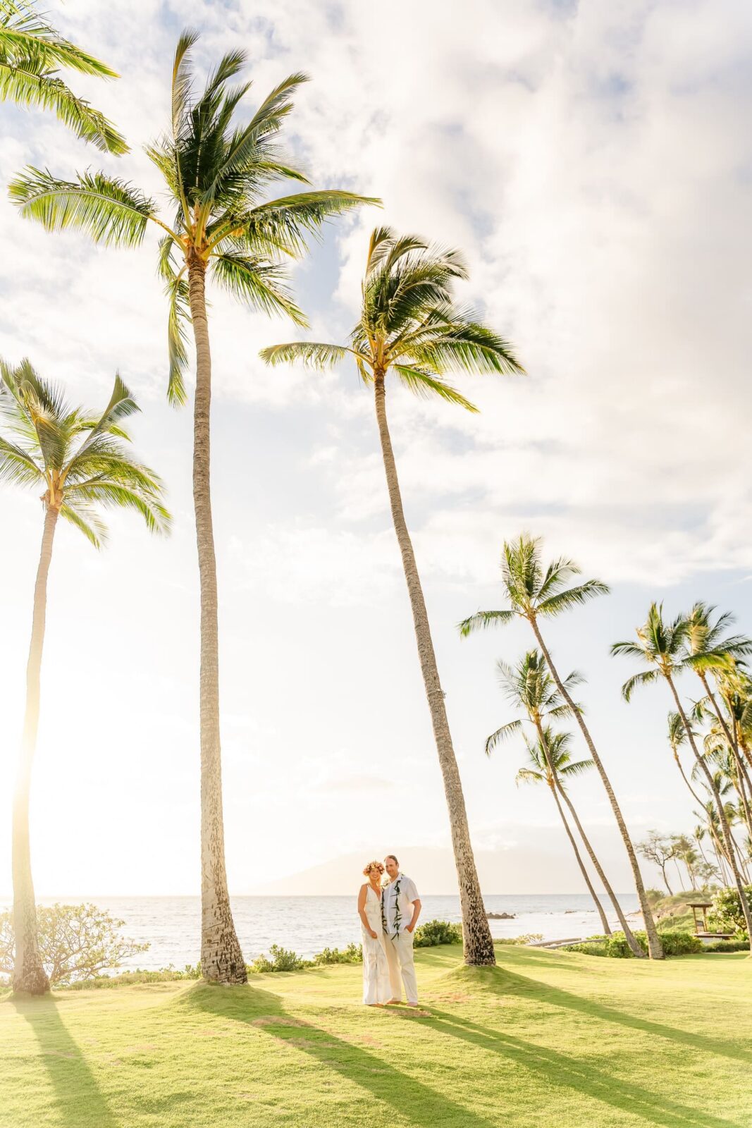 A couple in wedding attire stands on a grassy area by the ocean, surrounded by tall palm trees, with sunlight streaming through and a bright sky overhead.