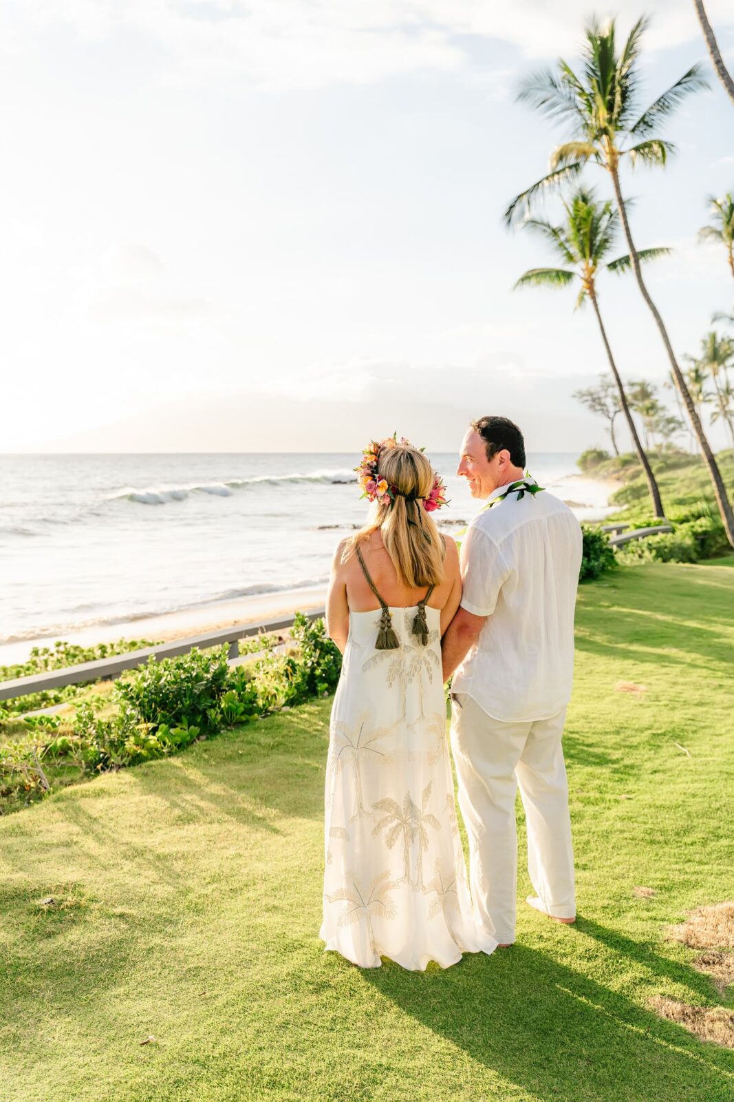 A couple dressed in white, with floral leis, stands on a grassy area by the ocean, looking at the waves. Palm trees and a sunny sky frame the tropical beach scene.