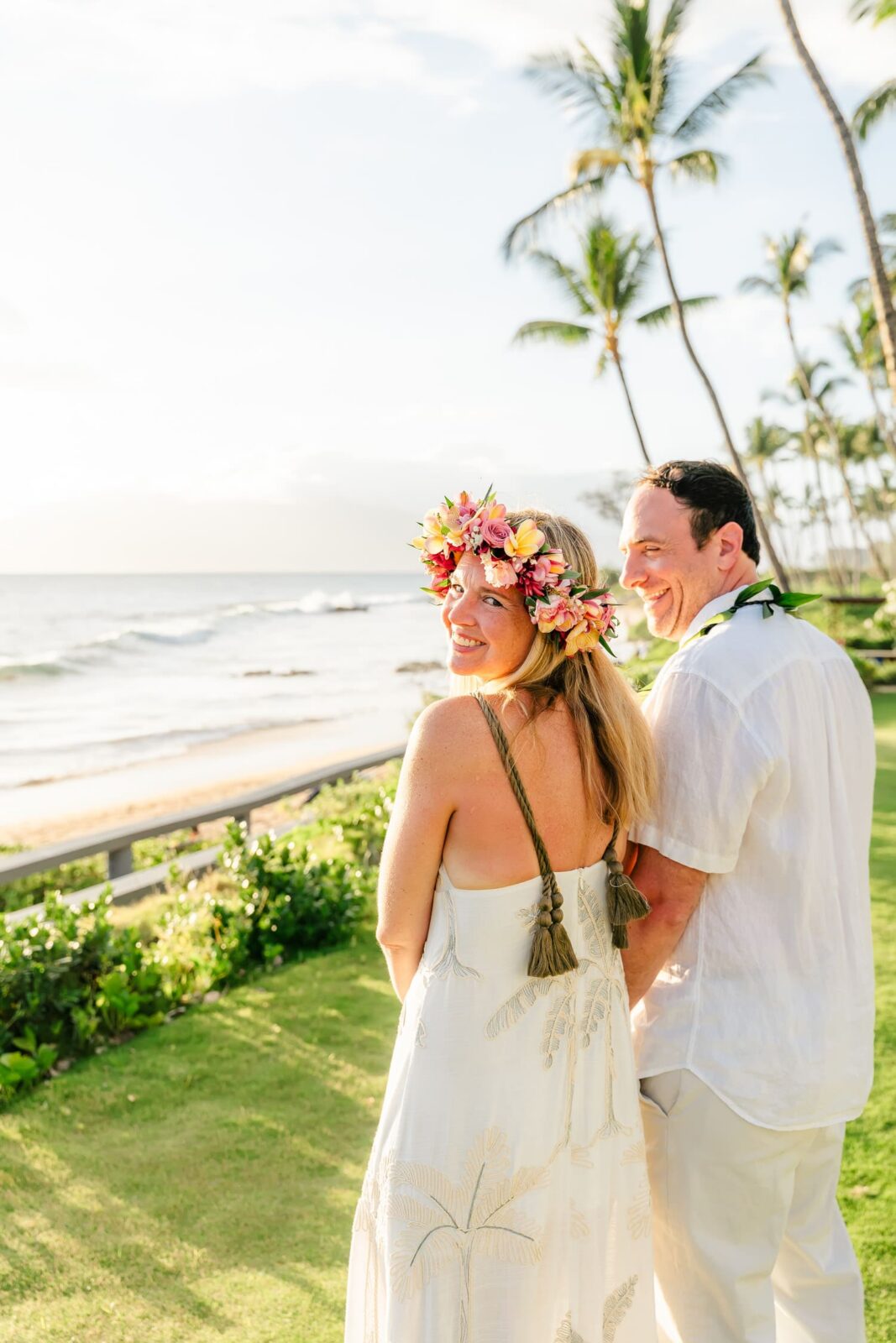 A smiling couple in white attire stands on a sunny beachside lawn; the woman wears a flower crown. Palm trees and ocean waves are visible in the background.