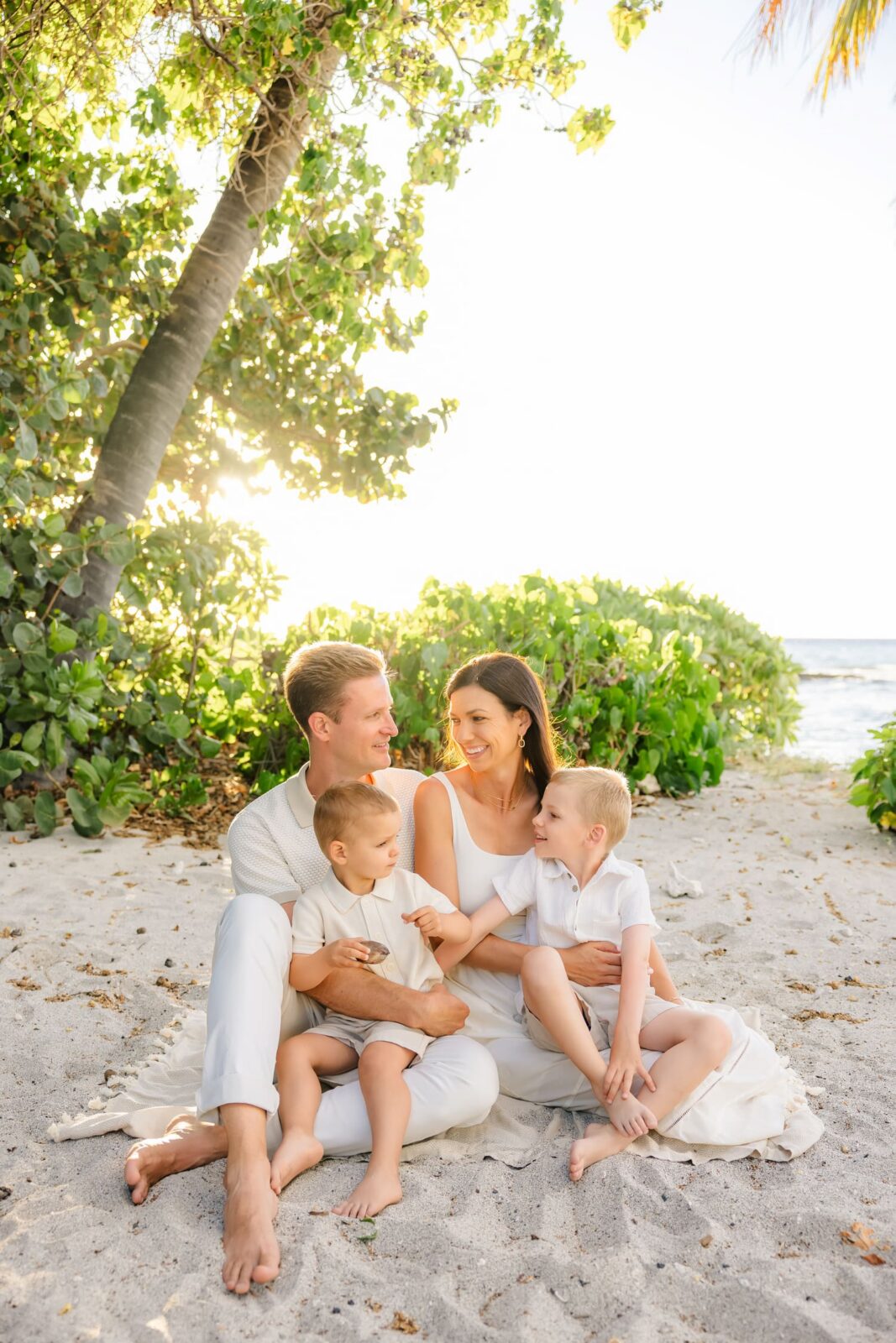 A family of four, dressed in white, sits smiling together on a sandy beach under green trees with sunlight filtering through, near the ocean.