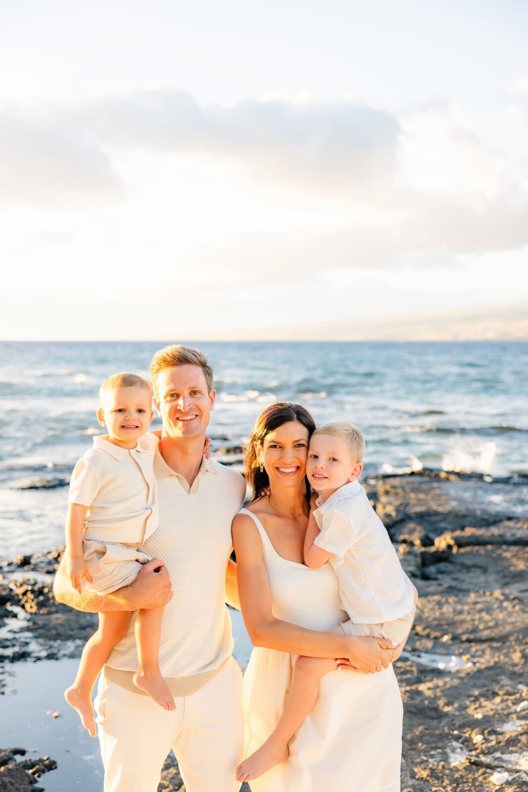 A smiling family of four poses on a beach at sunset, with the parents each holding a young child. The family wears light-colored clothing, and the ocean and sky are visible in the background.