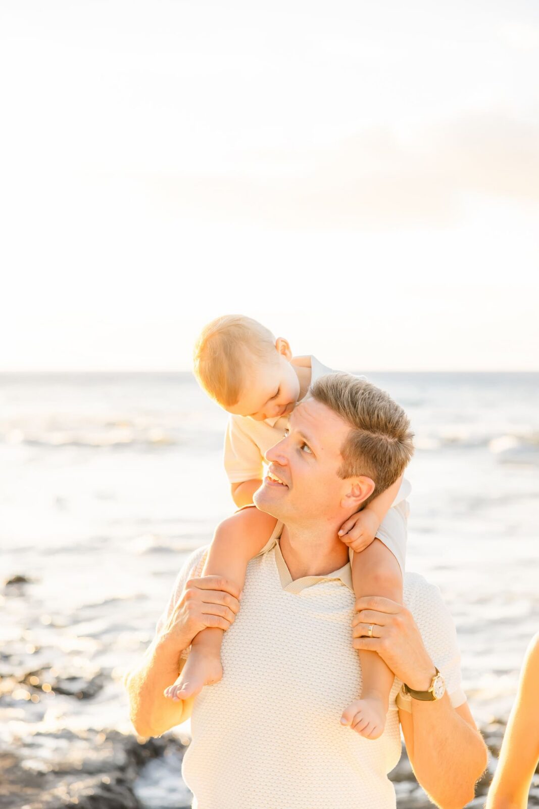 A man smiles while carrying a young child on his shoulders at the beach, with the ocean and bright sky in the background.