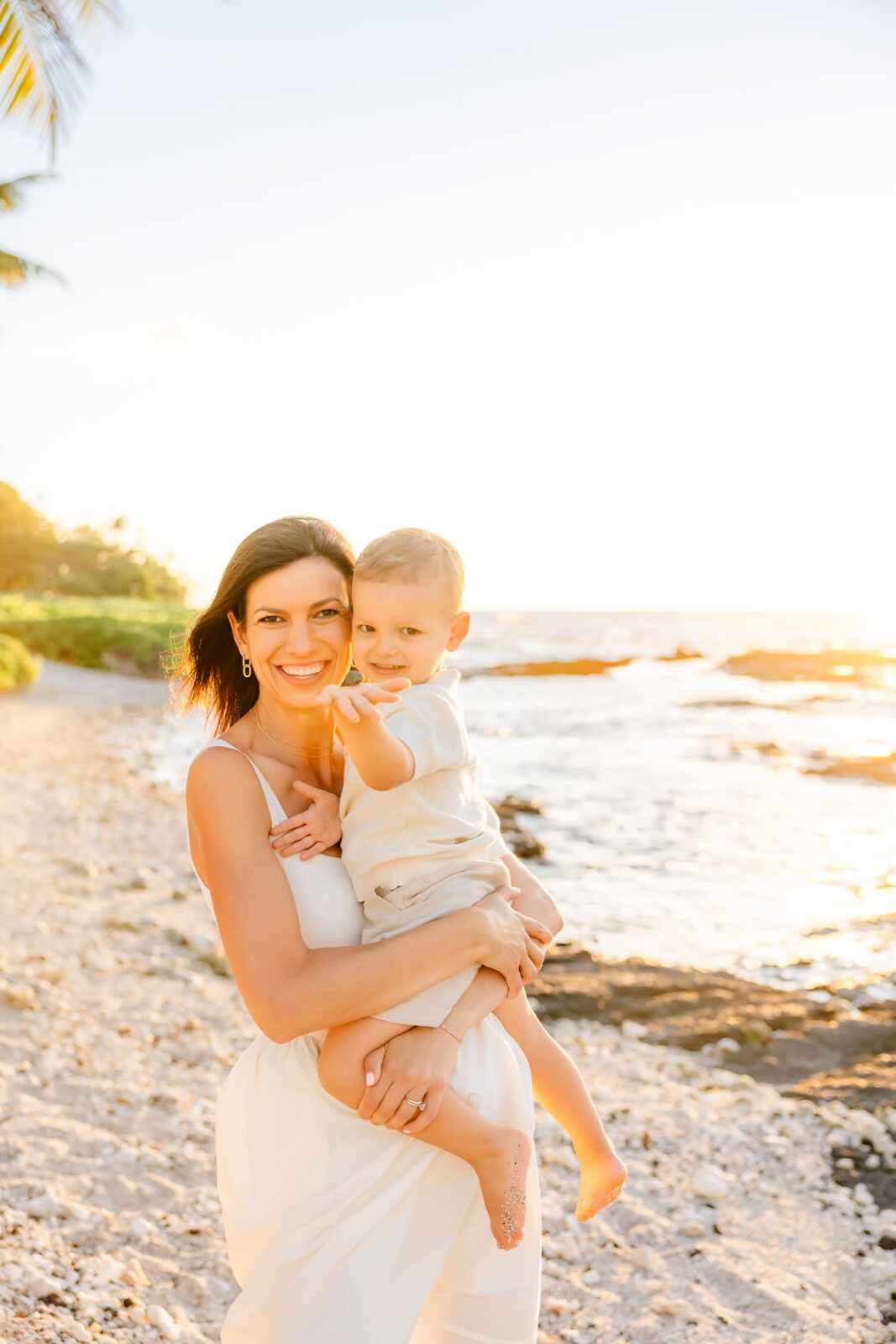 A woman in a white dress smiles while holding a young child on a rocky beach at sunset. The ocean and bright sky are in the background.