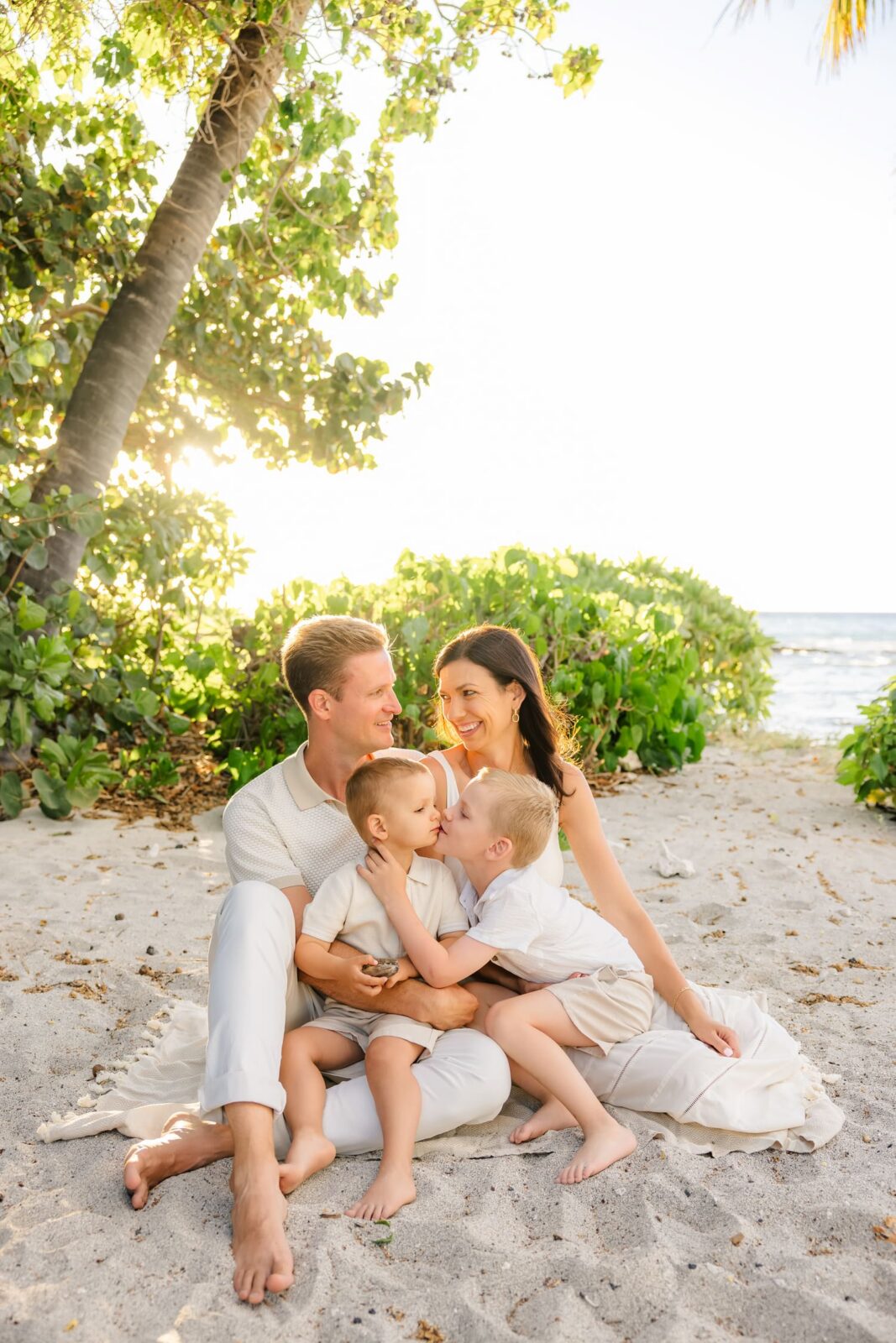 A family of four sits closely together on a sandy beach, smiling and laughing as sunlight filters through trees and greenery behind them, with the ocean in the background.