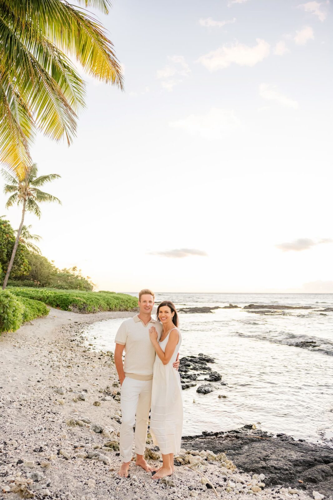 A couple in white outfits stands barefoot on a rocky beach at sunset, with palm trees and greenery in the background and gentle waves by their feet.