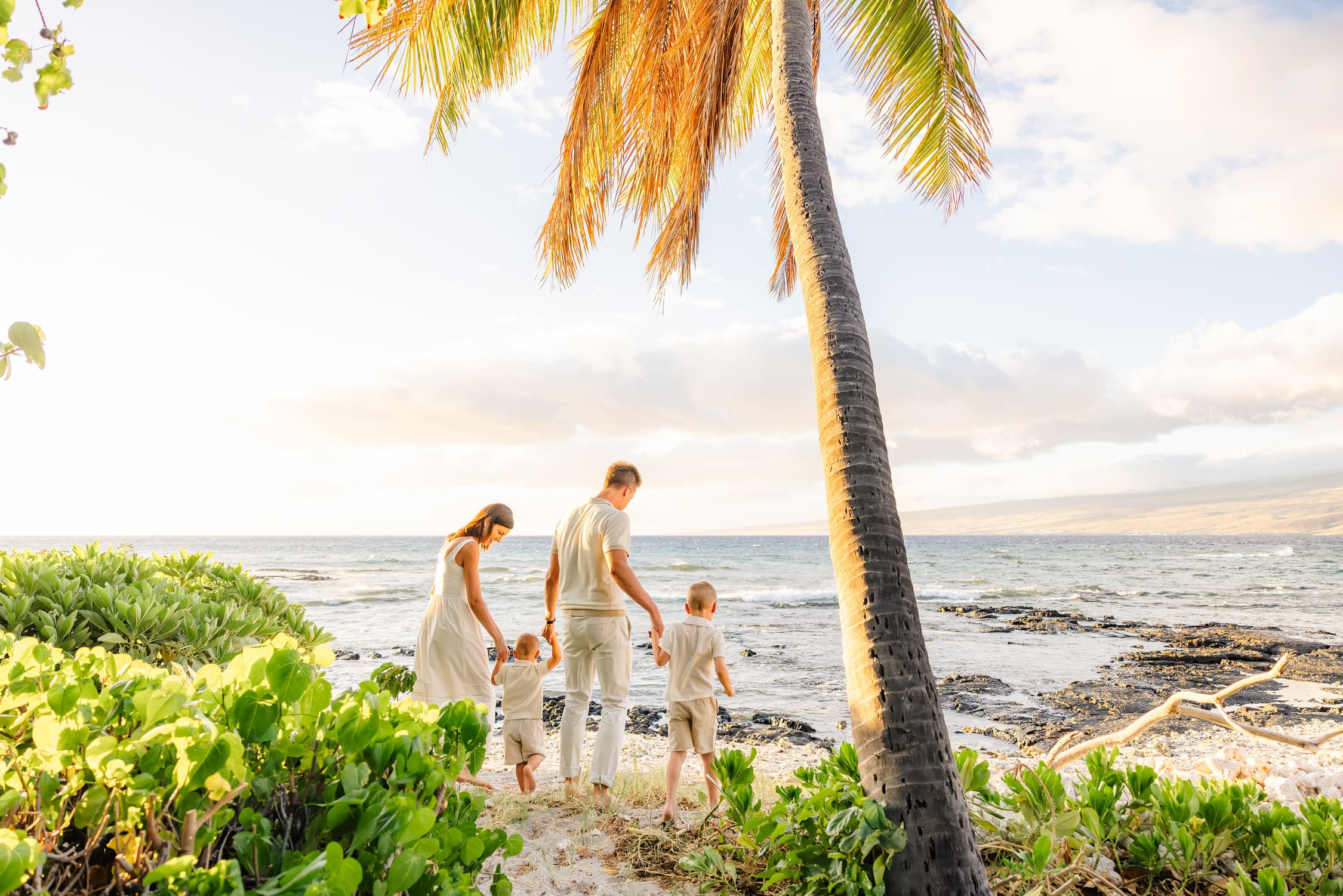 A family of four, dressed in light colors, walks along a sandy beach under a palm tree, with ocean waves and a bright sky in the background.