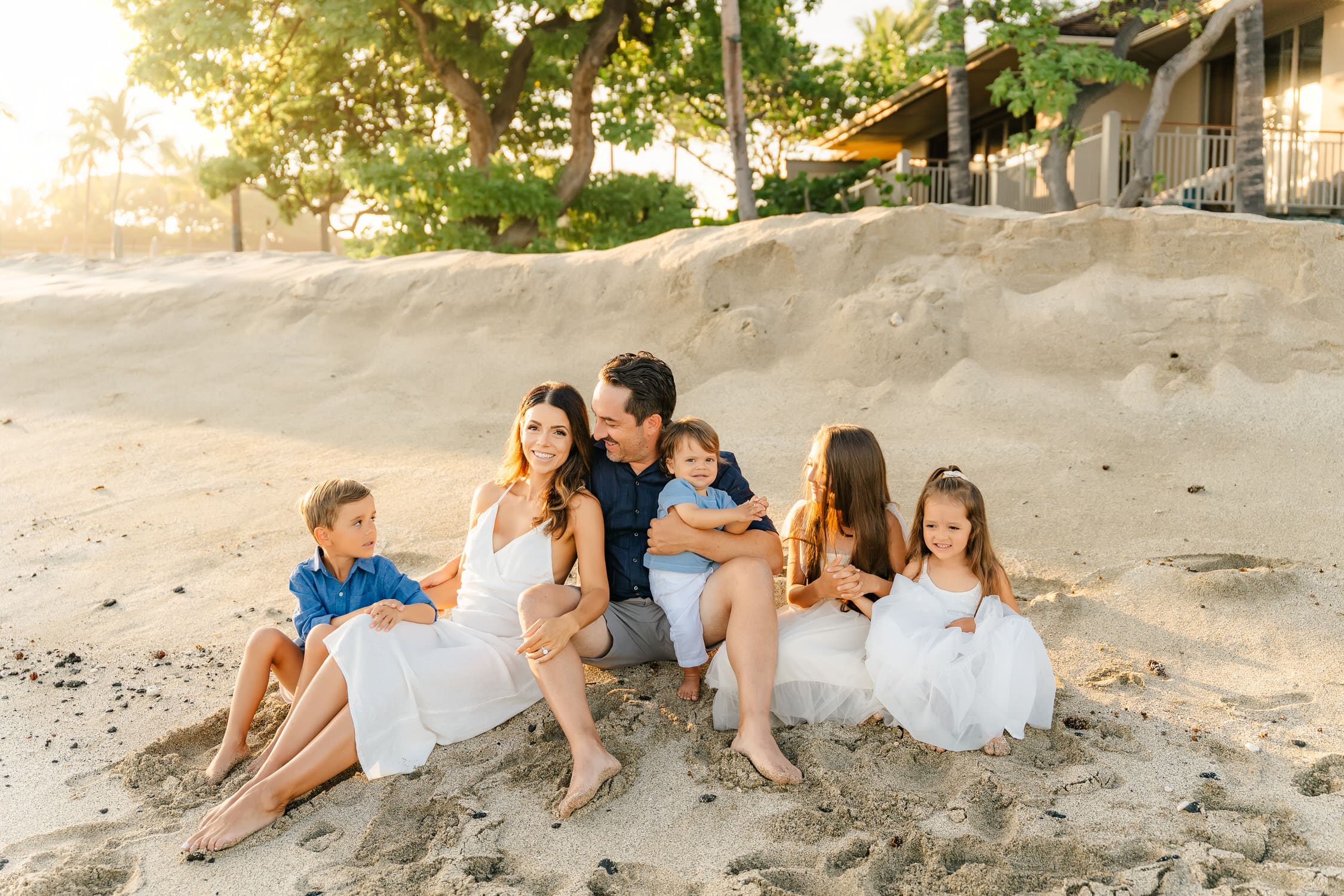 A family of six sits together on a sandy beach at sunset, smiling and dressed in white and blue outfits, with trees and a house behind them—a perfect moment beautifully captured by a Four Seasons Hualalai family photographer.