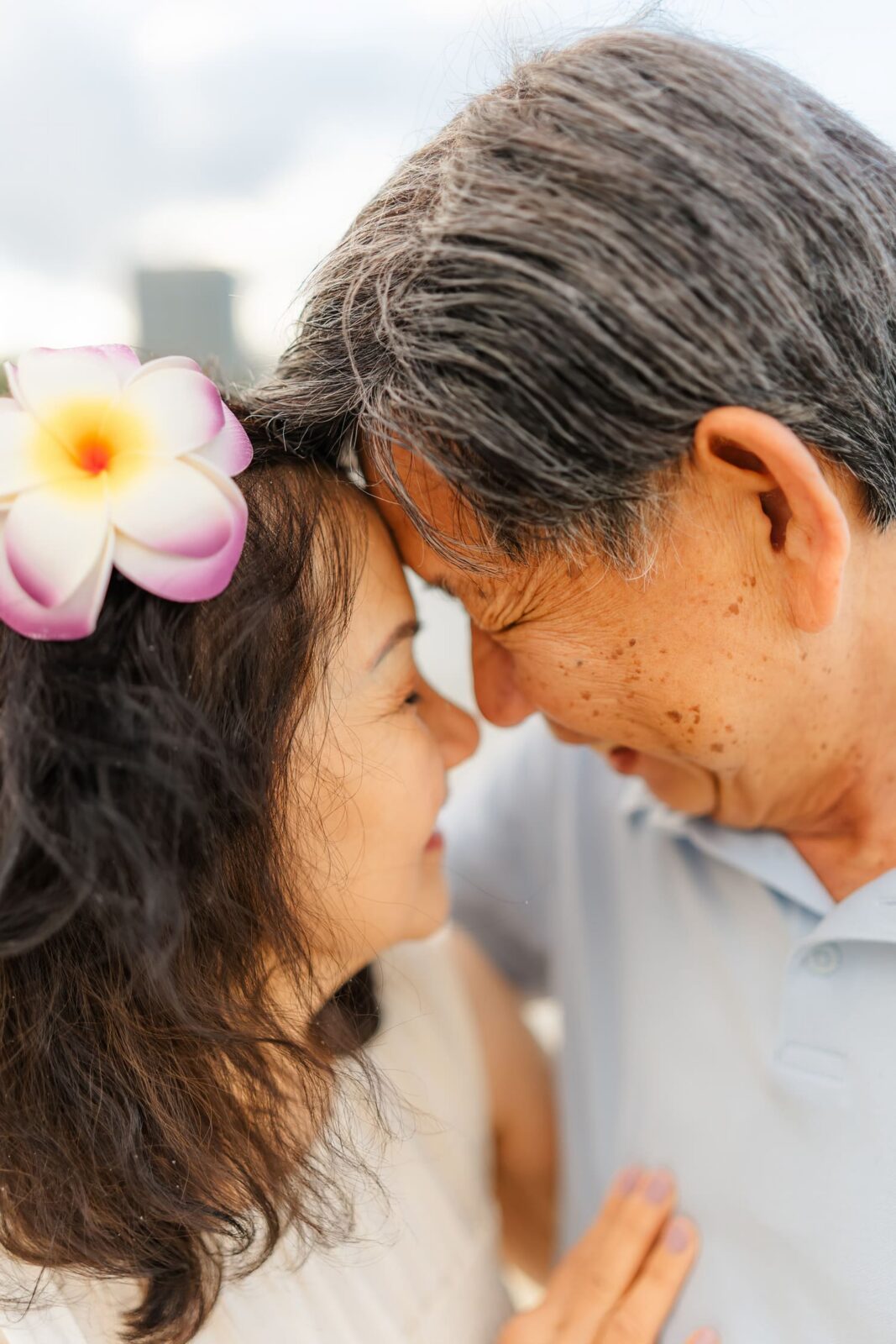 An older couple, smiling with their foreheads touching, shares a tender moment. The woman wears a flower in her hair. Both appear joyful and close, with a soft, blurred background.