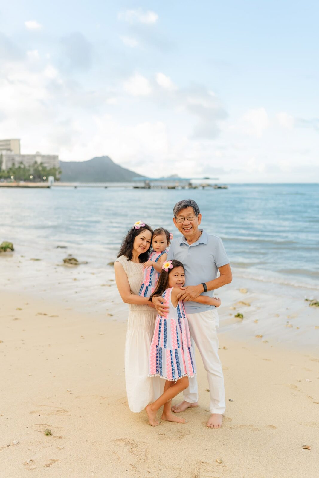 A smiling family of four poses on a sandy beach with mountains and ocean in the background. The two young girls wear matching dresses. The mother and father stand close, embracing their daughters.