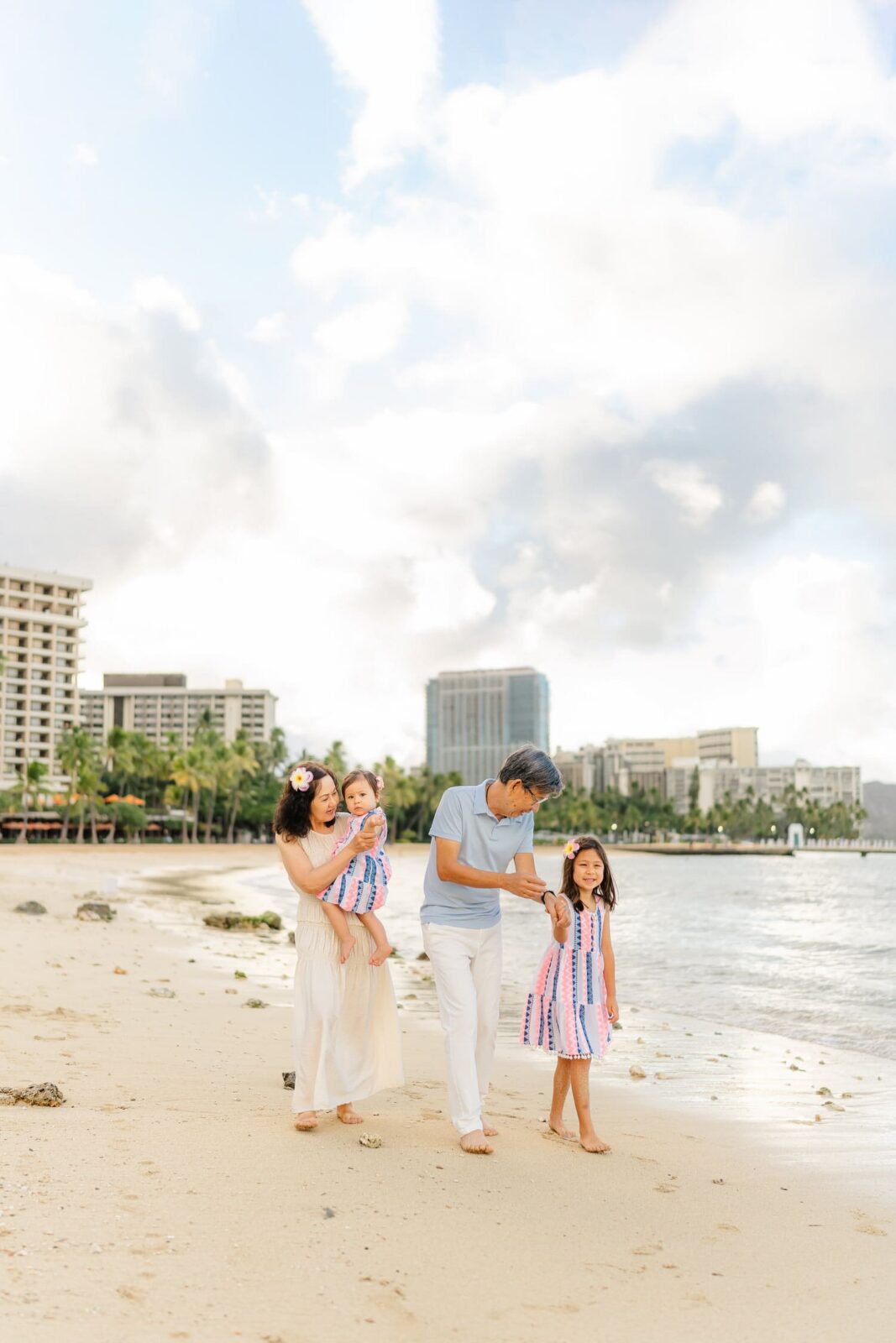 A family of four walks barefoot on a sandy beach near the water, with city buildings and palm trees in the background under a cloudy sky.
