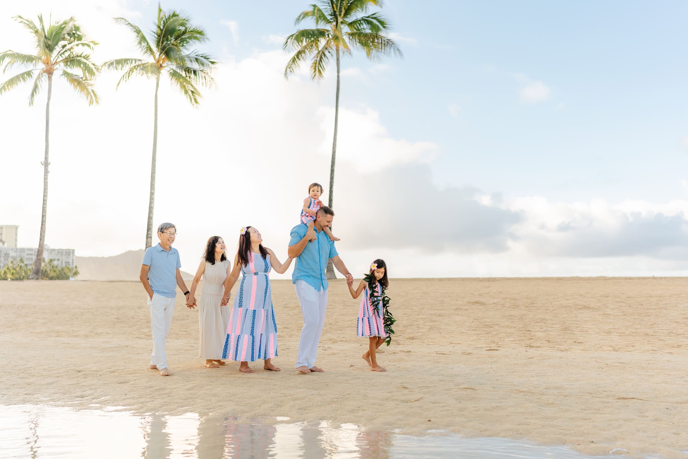 A family of six walks hand-in-hand on a sandy beach, with palm trees and blue sky in the background. One child sits on an adults shoulders, and everyone looks happy and relaxed by the water.
