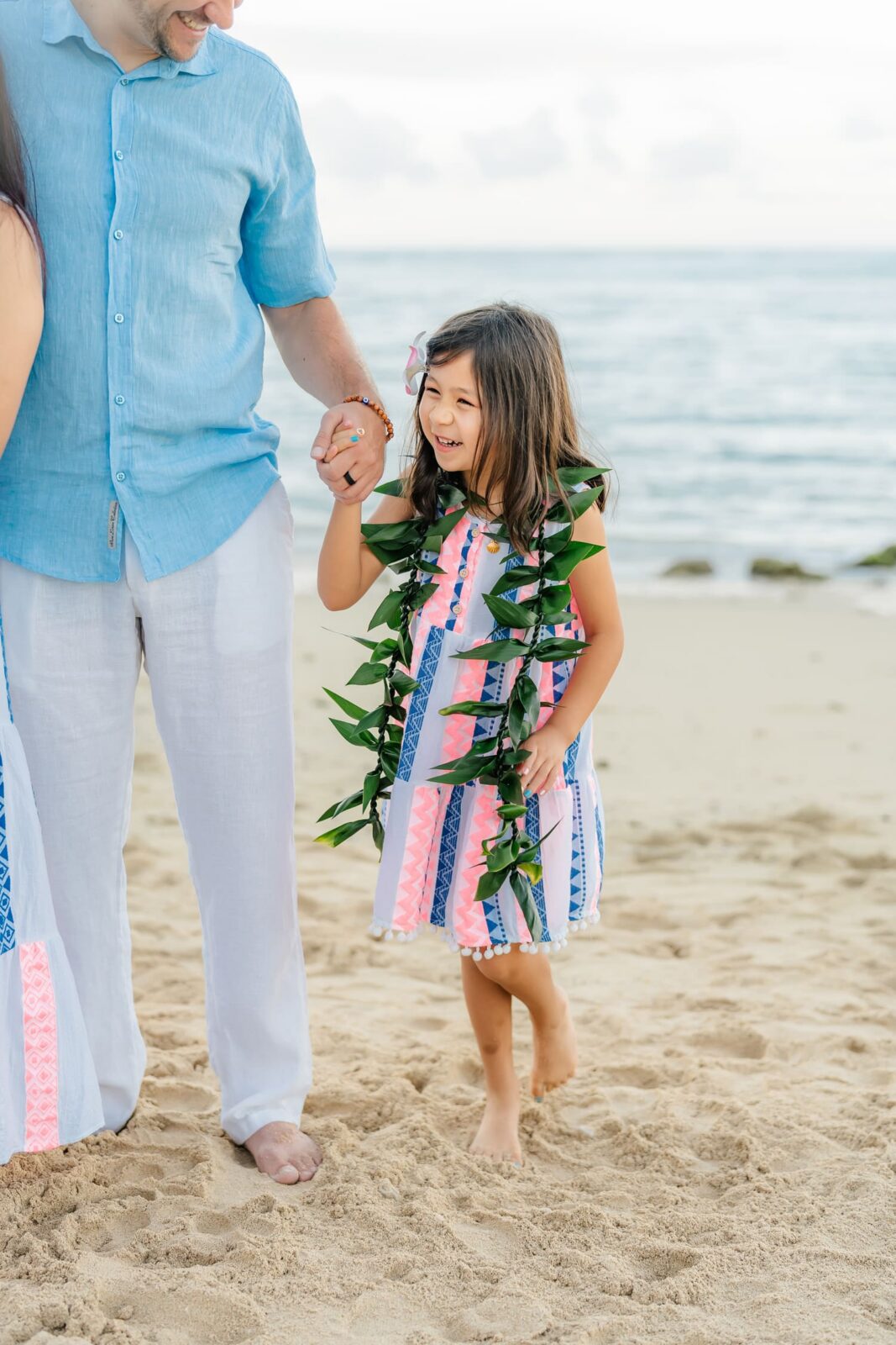 Smiling young girl in a colorful dress and leafy lei holds an adults hand while walking barefoot on a sandy beach near the ocean.