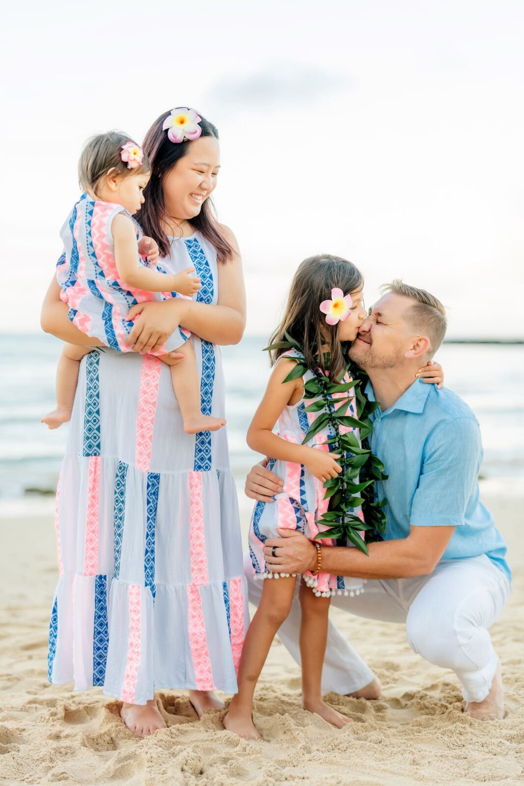 A family of four stands on a sandy beach. The mother holds a young girl, while the father kneels, kissing an older daughter. Everyone wears summery, patterned outfits and flower hair clips. The ocean and sky are in the background.
