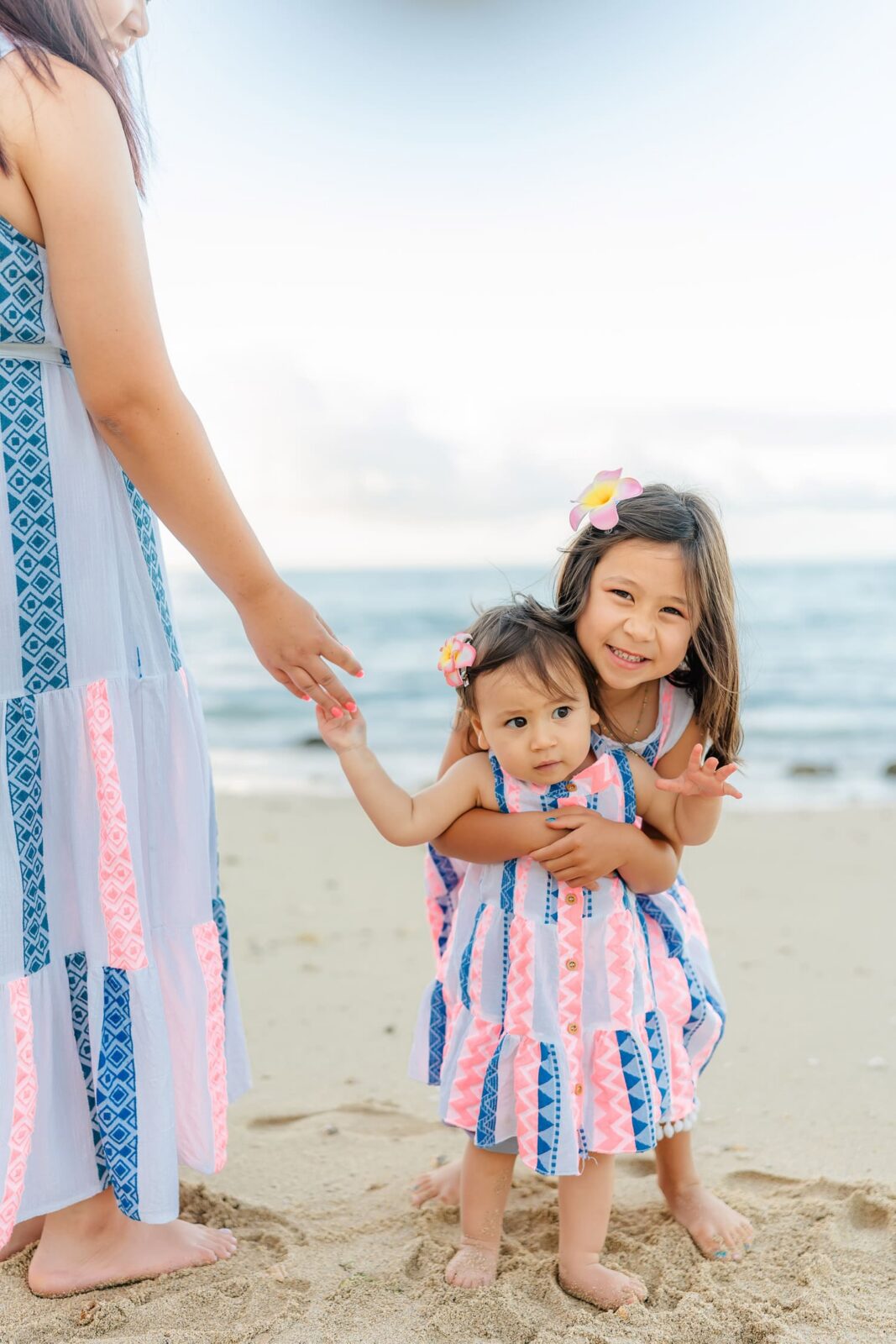 Two young girls in matching blue and pink dresses stand on a beach, one hugging the other. An adult wearing a similar dress extends a hand toward them. The ocean and sky are in the background.