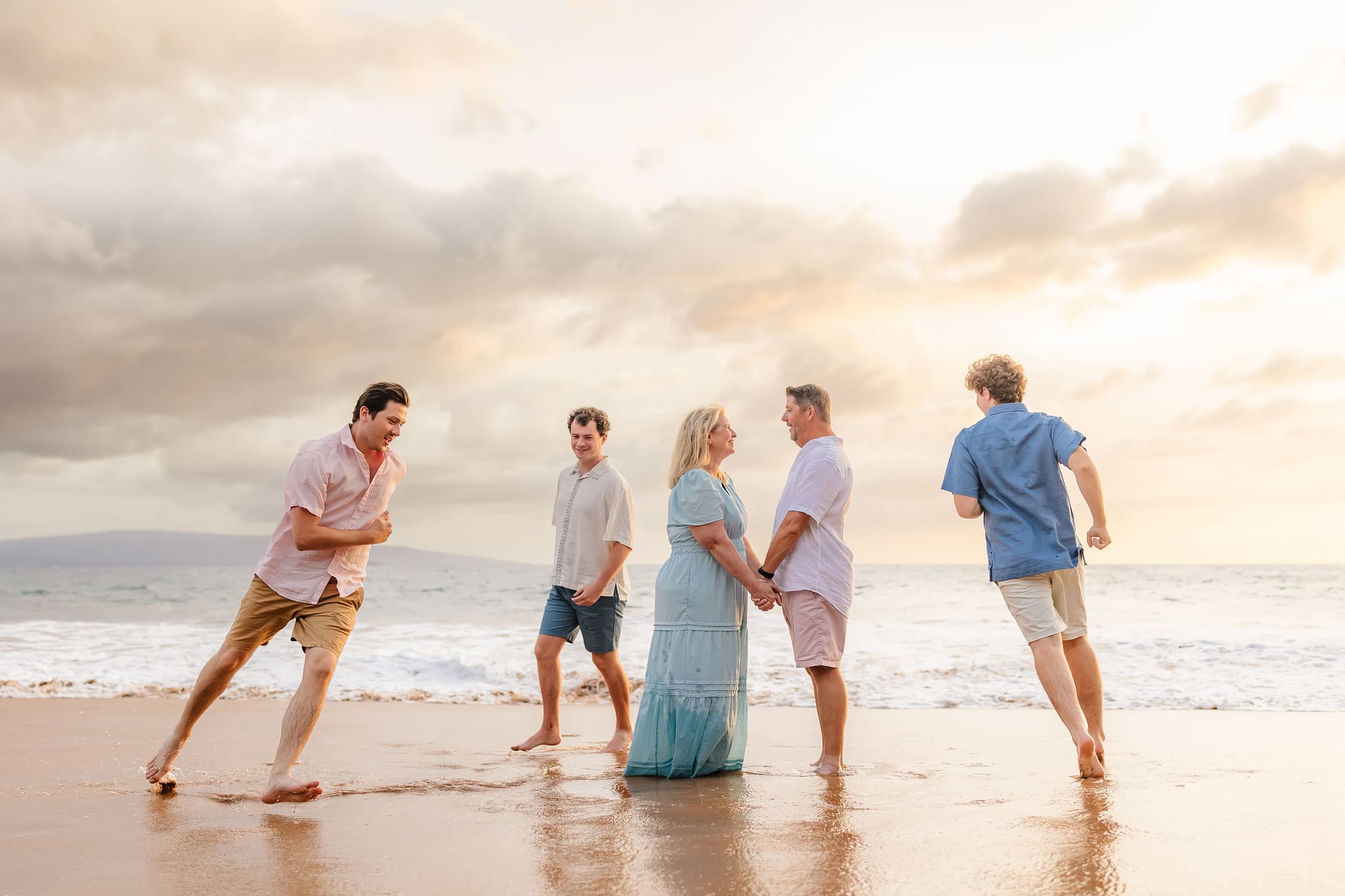A family of five stands barefoot on a sandy beach at sunset. The parents hold hands and face each other while three young men walk and run nearby. The ocean and cloudy sky are in the background.
