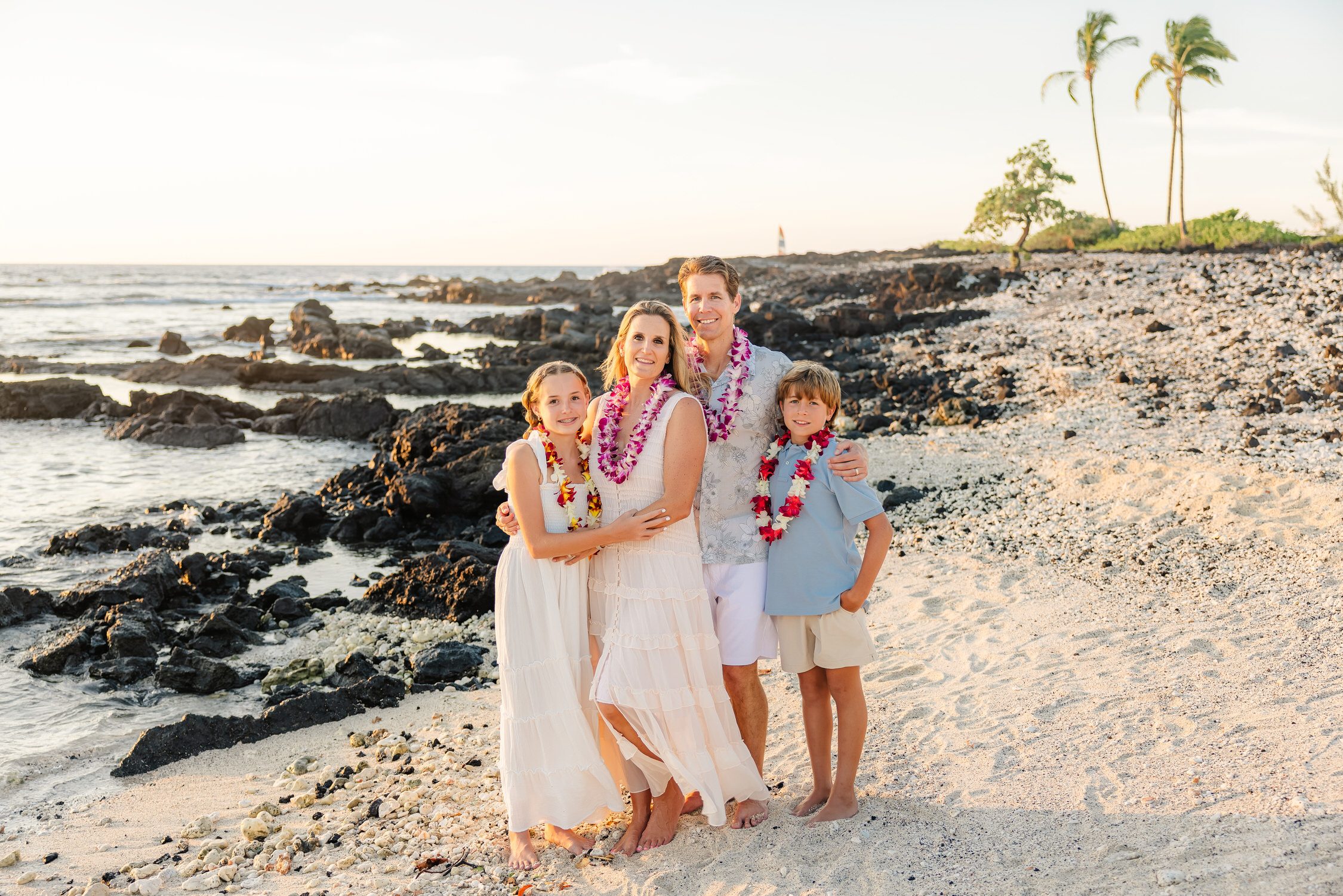 Family of four in white and soft blue, wearing fuchsia and orchid lei, standing together on a white sand and lava rock shoreline near Anaehoomalu Bay at sunset.
