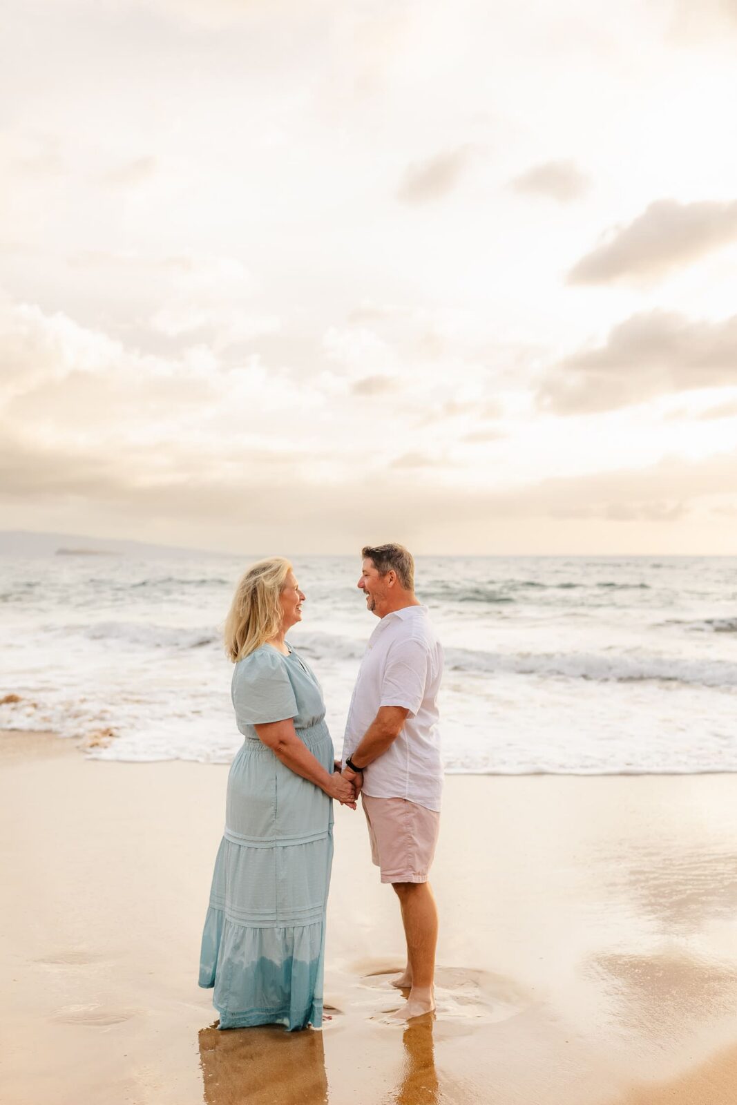 A couple stands barefoot on a sandy beach at sunset, holding hands and smiling at each other with waves and a cloudy sky in the background—capturing the kind of heartfelt moment a Wailea family photographer would treasure.